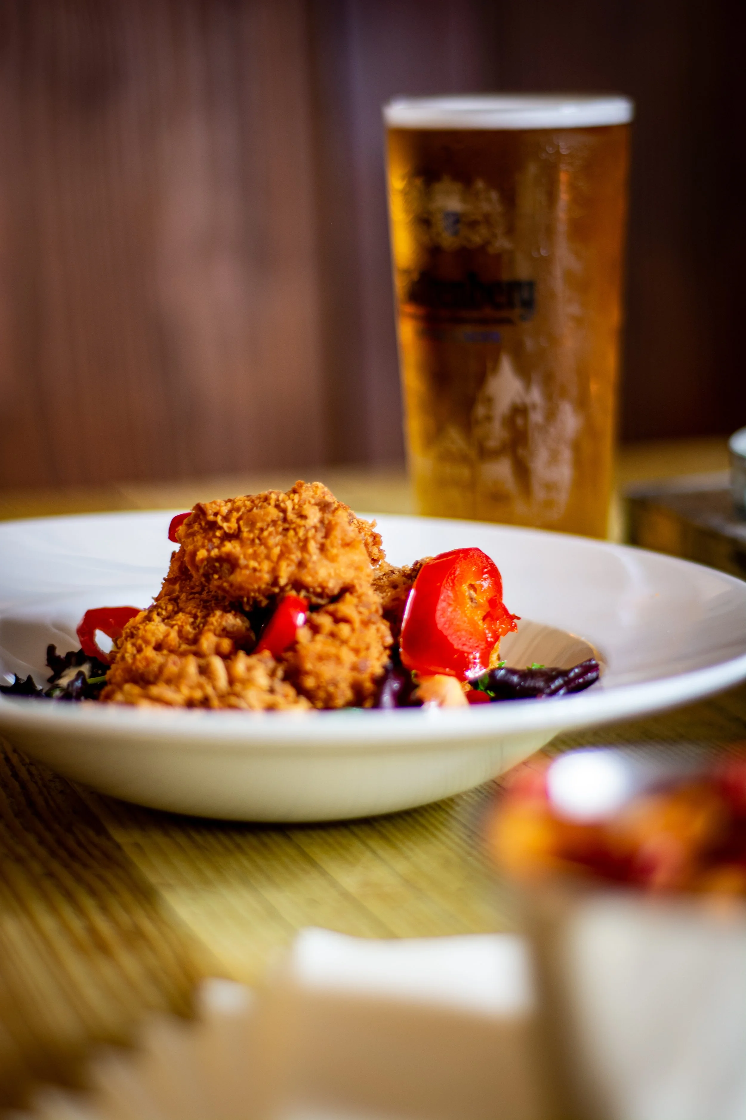 Fried chicken bites with chillis and greens on a white plate, with a glass of beer in the background.