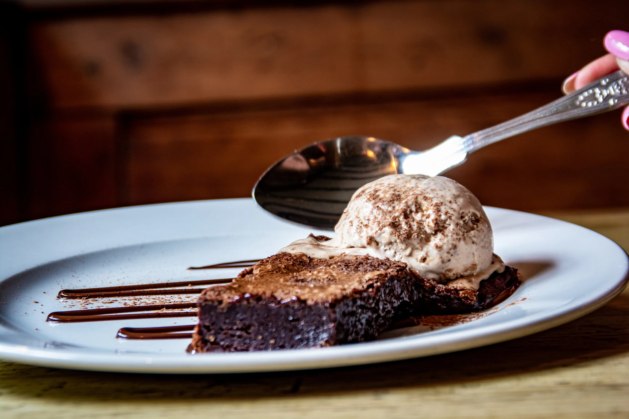 Chocolate brownie with a scoop of vanilla ice cream sprinkled with cocoa powder, drizzled with chocolate sauce on a white plate being served with a spoon.