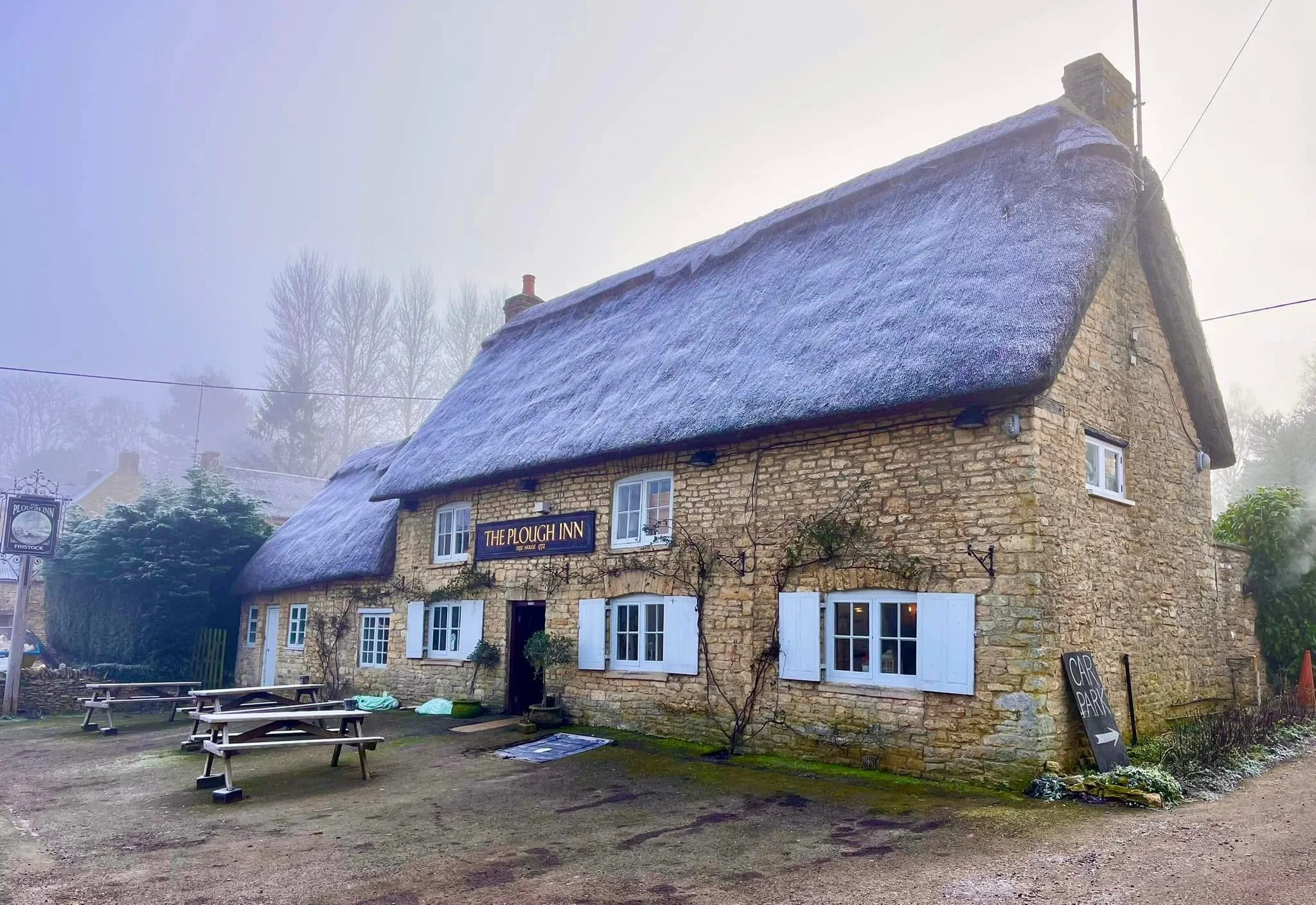 A stone building with a thatched roof and white window shutters, labeled 'The Plough Inn', with outdoor picnic tables and a sign for car parking, on a foggy day.