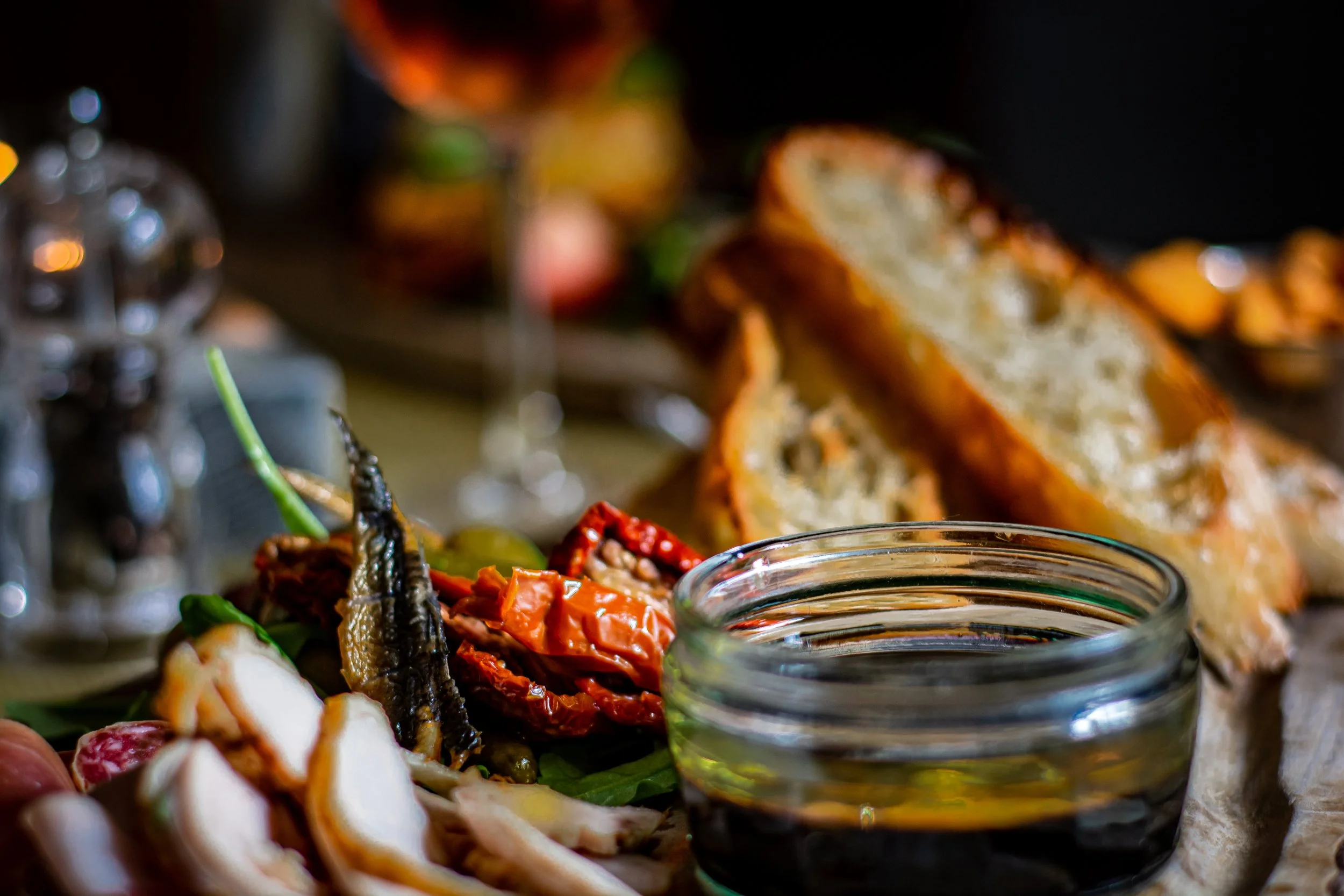 Close-up of a rustic picnic spread with sliced bread, sun-dried tomatoes, grilled vegetables, a glass bowl of olive oil and balsamic vinegar, and a bowl of olives on a wooden table.