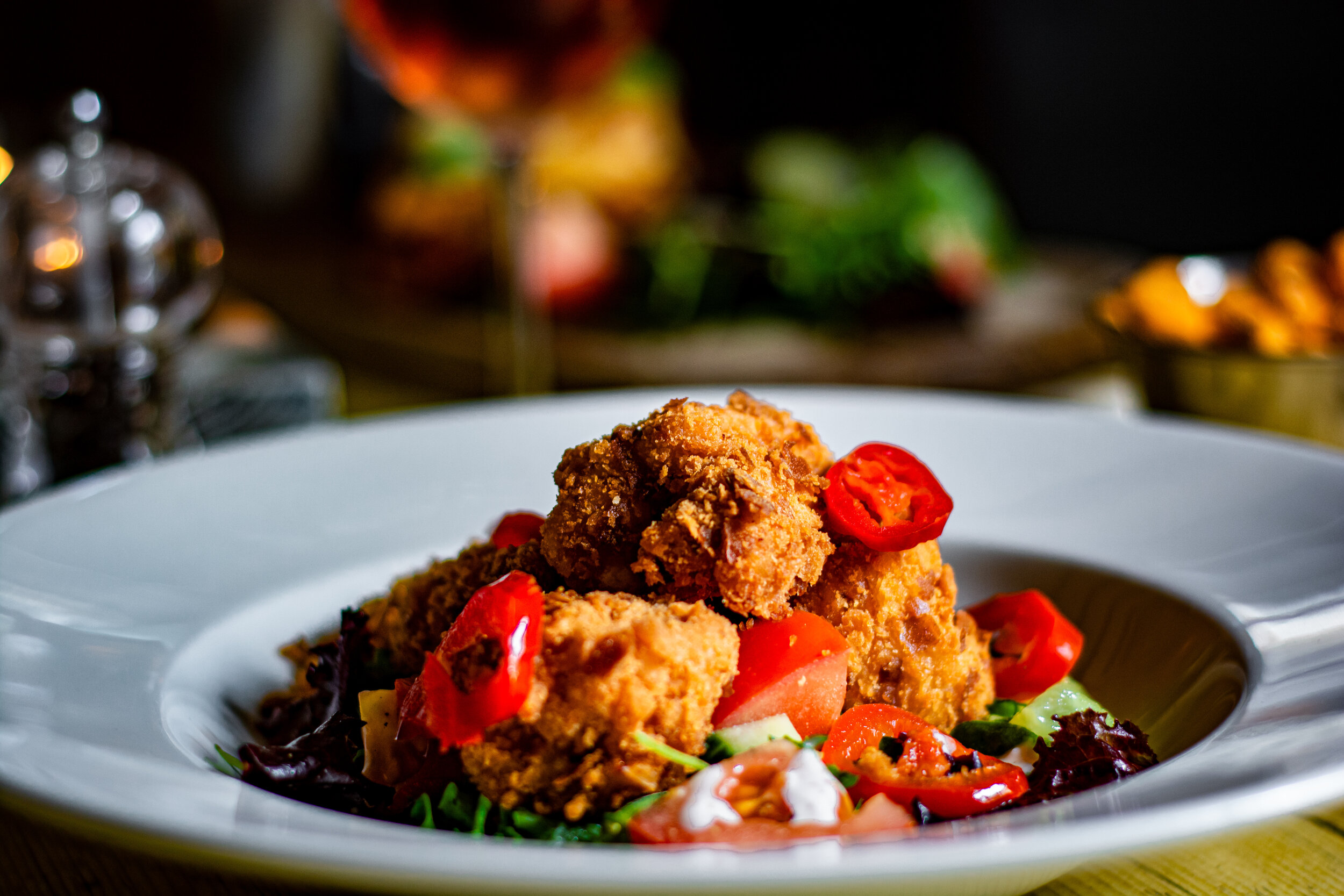 Fried chicken pieces served on a bed of salad with cucumbers, and red chili peppers on a white plate.