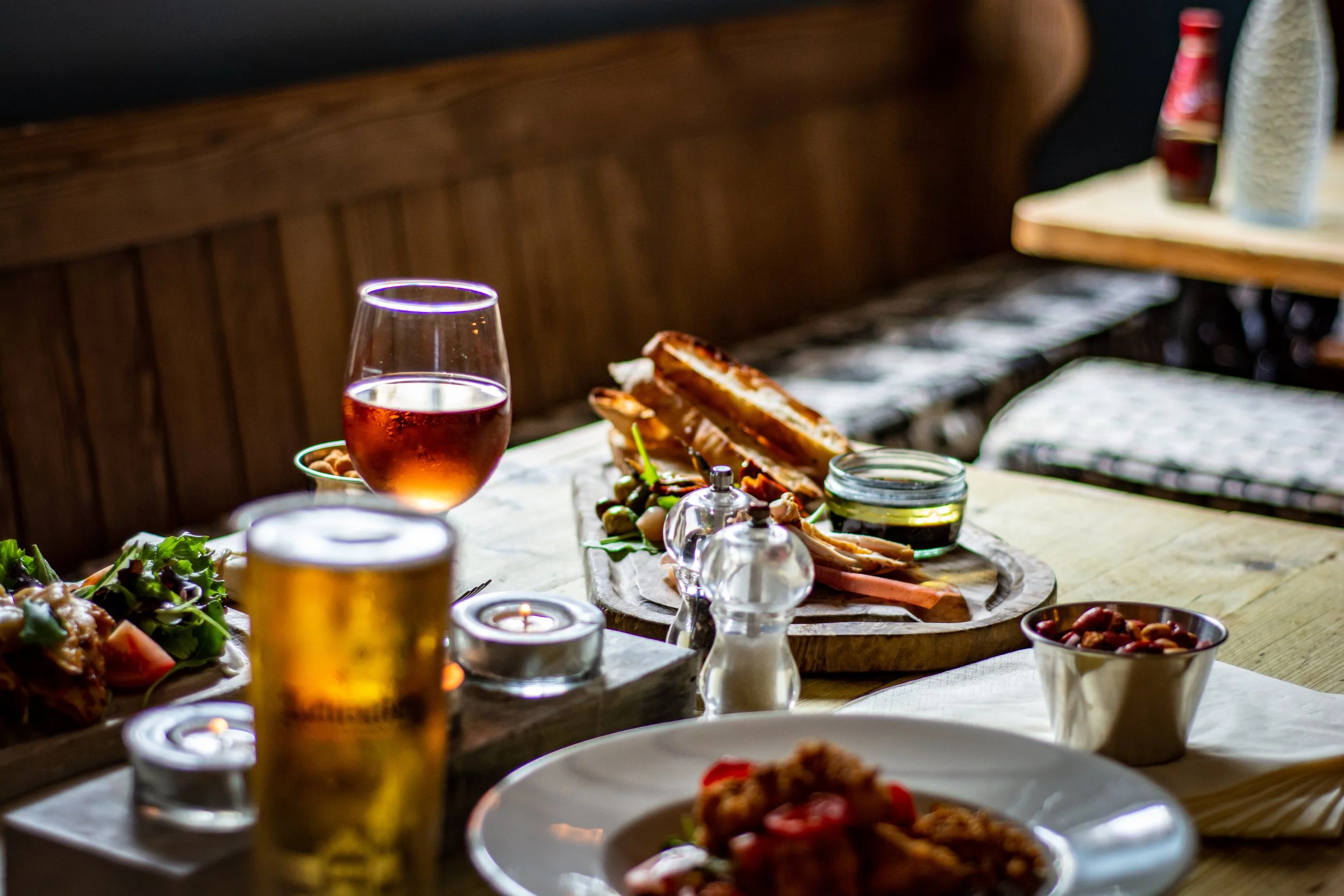 A table set with various food and drinks including a glass of rosé wine, a beer, a bowl of nuts, a salad, a sandwich on a wooden serving board, a small bowl of pickles, and a bowl of mixed nuts, with a cozy restaurant or cafe setting.