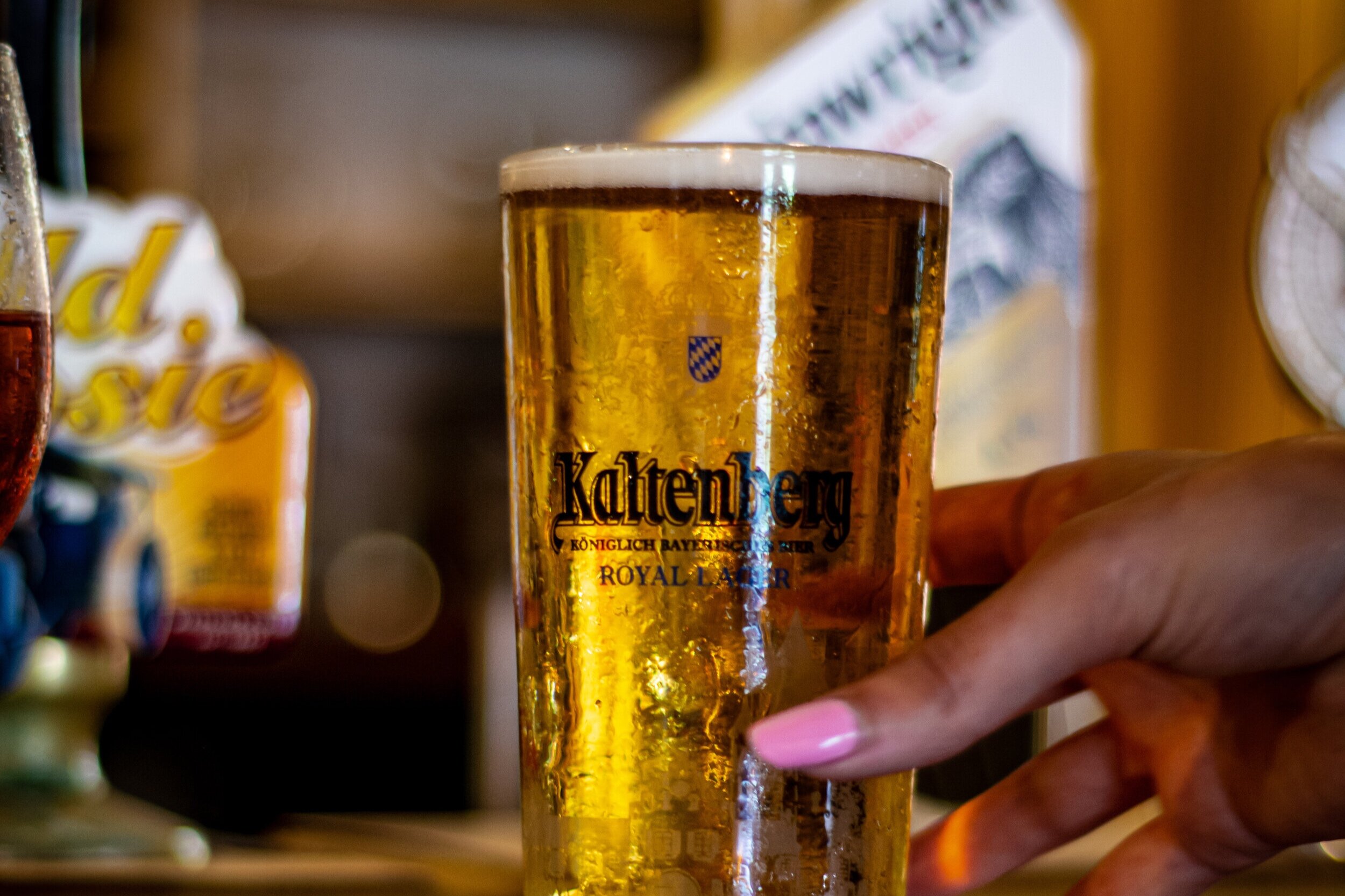 A hand holding a tall glass of beer on a bar counter, with a blurred background of beer taps and bar signs.