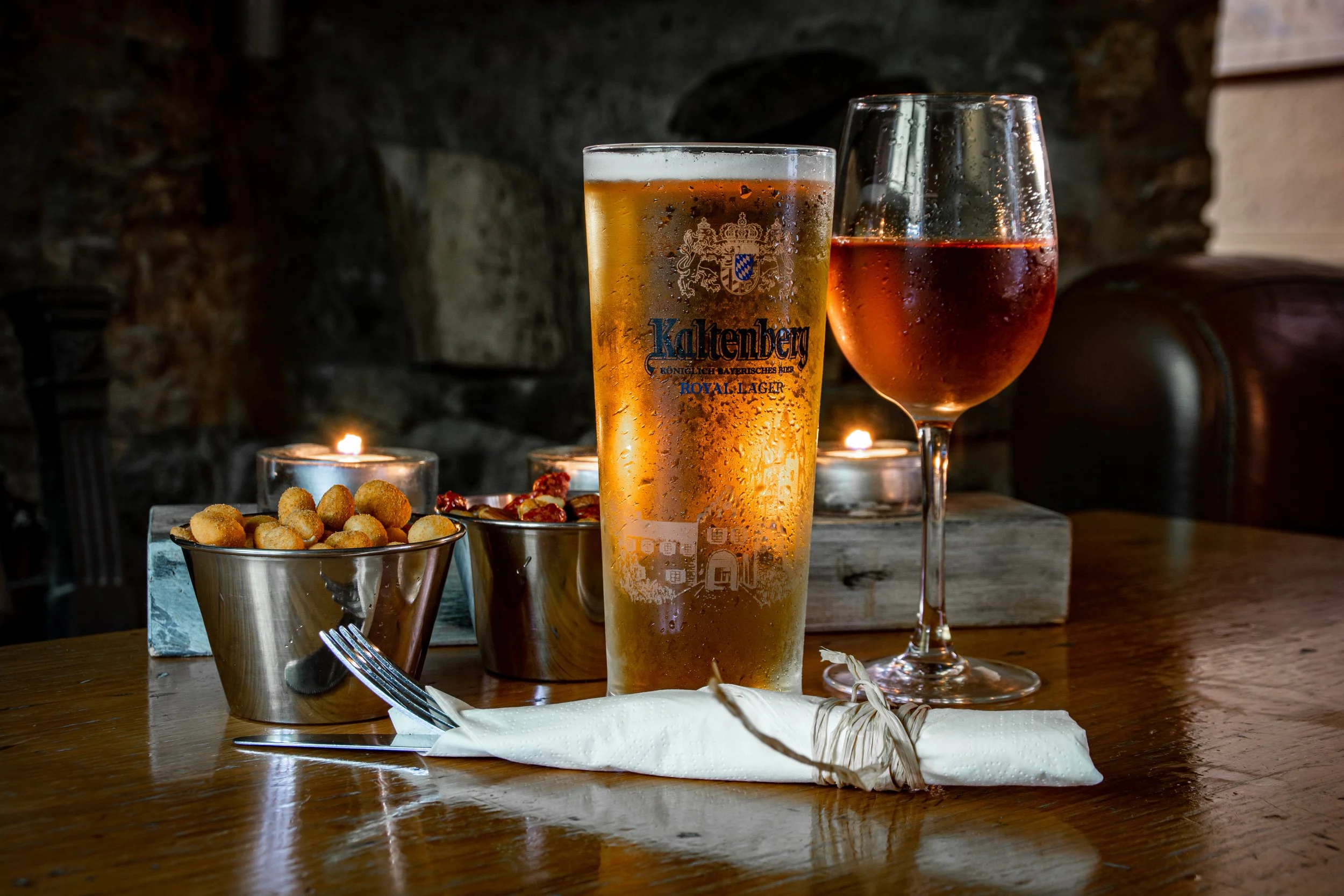 A glass of beer and a glass of rosé wine on a table, with two small bowls of snacks, lit candles, and a napkin with a fork and knife in a cozy restaurant setting.