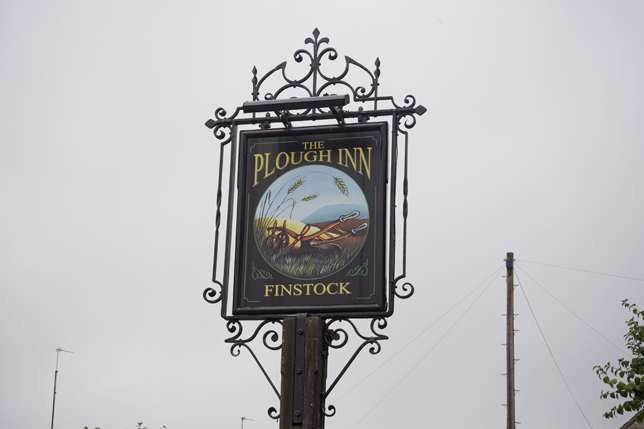 Photograph of a decorative hanging sign for The Plough Inn in Finstock, featuring an illustration of a plow and wheat in a circular frame on a cloudy day.