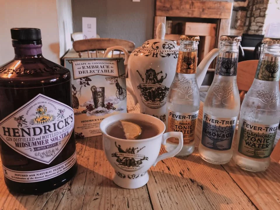 A wooden table with a bottle of Hendrick's gin, a cup of tea with lemon, three bottles of Fever-Tree tonic water, and a box of Hendrick's drink mix.