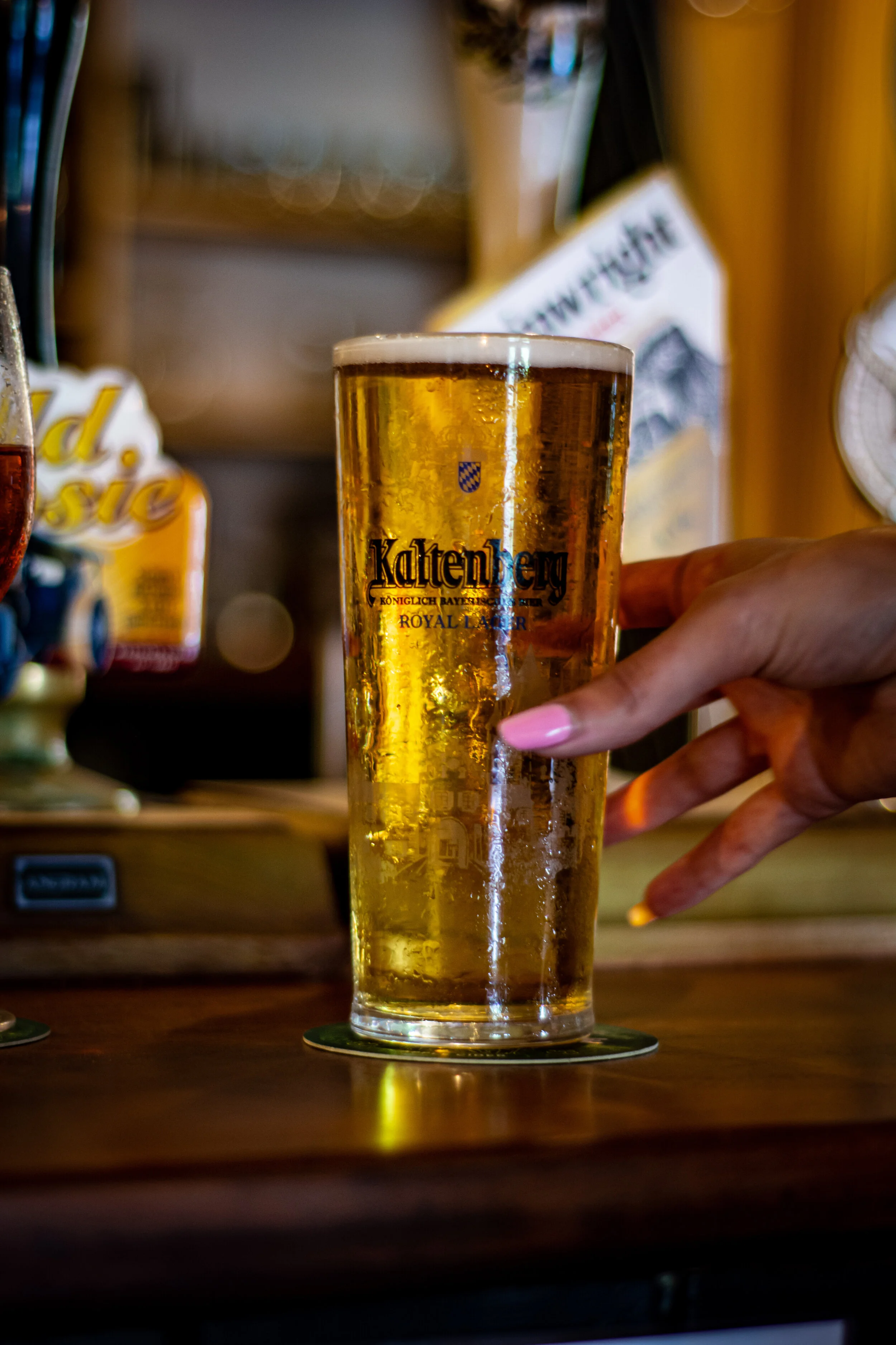 A tall glass of Karlsberg beer on a bar counter with a hand holding it, with a blurred background of bar decor.