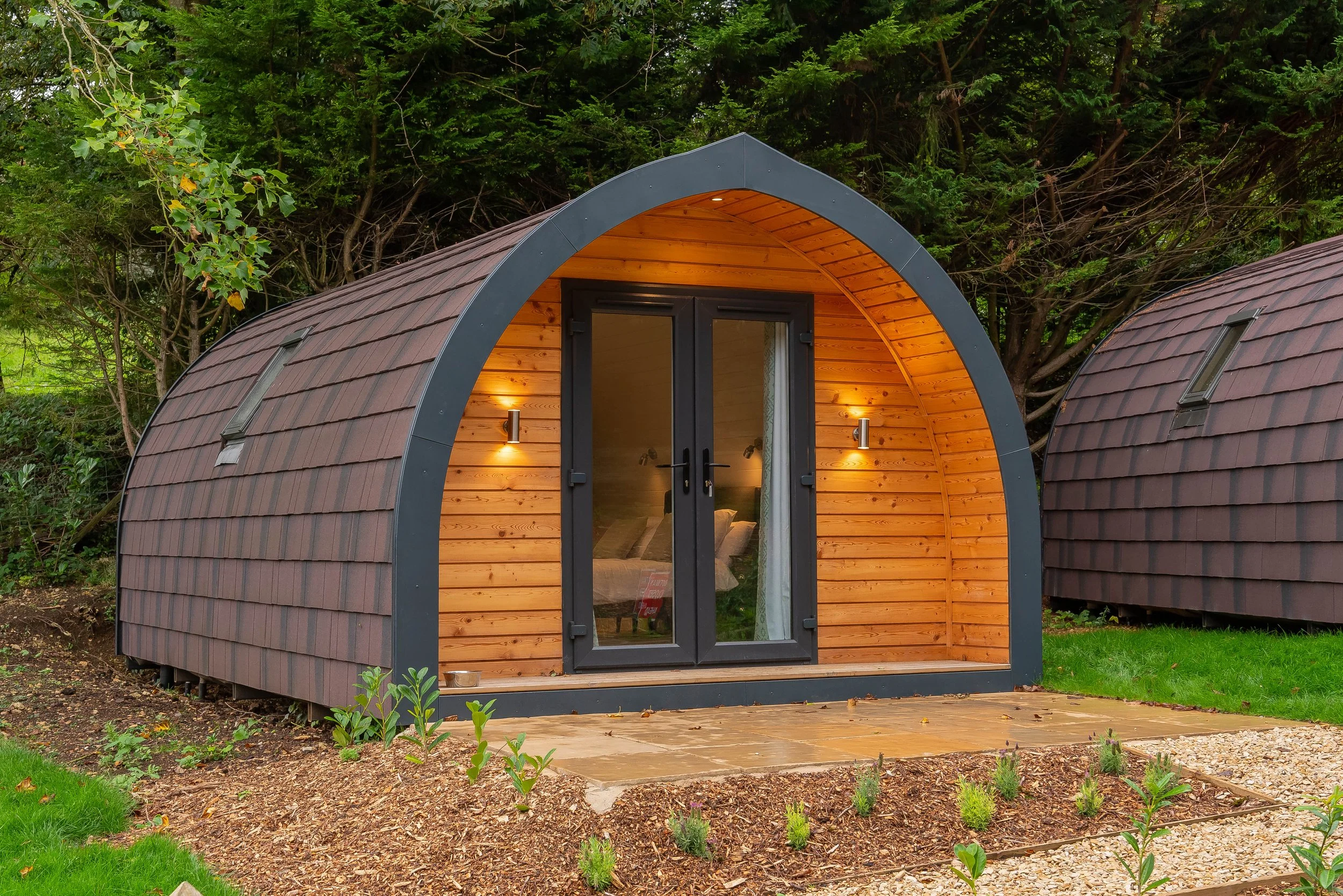 A modern round-shaped tiny house with a wooden interior, large glass double doors, and exterior with shingle siding, surrounded by green grass, plants, and trees.