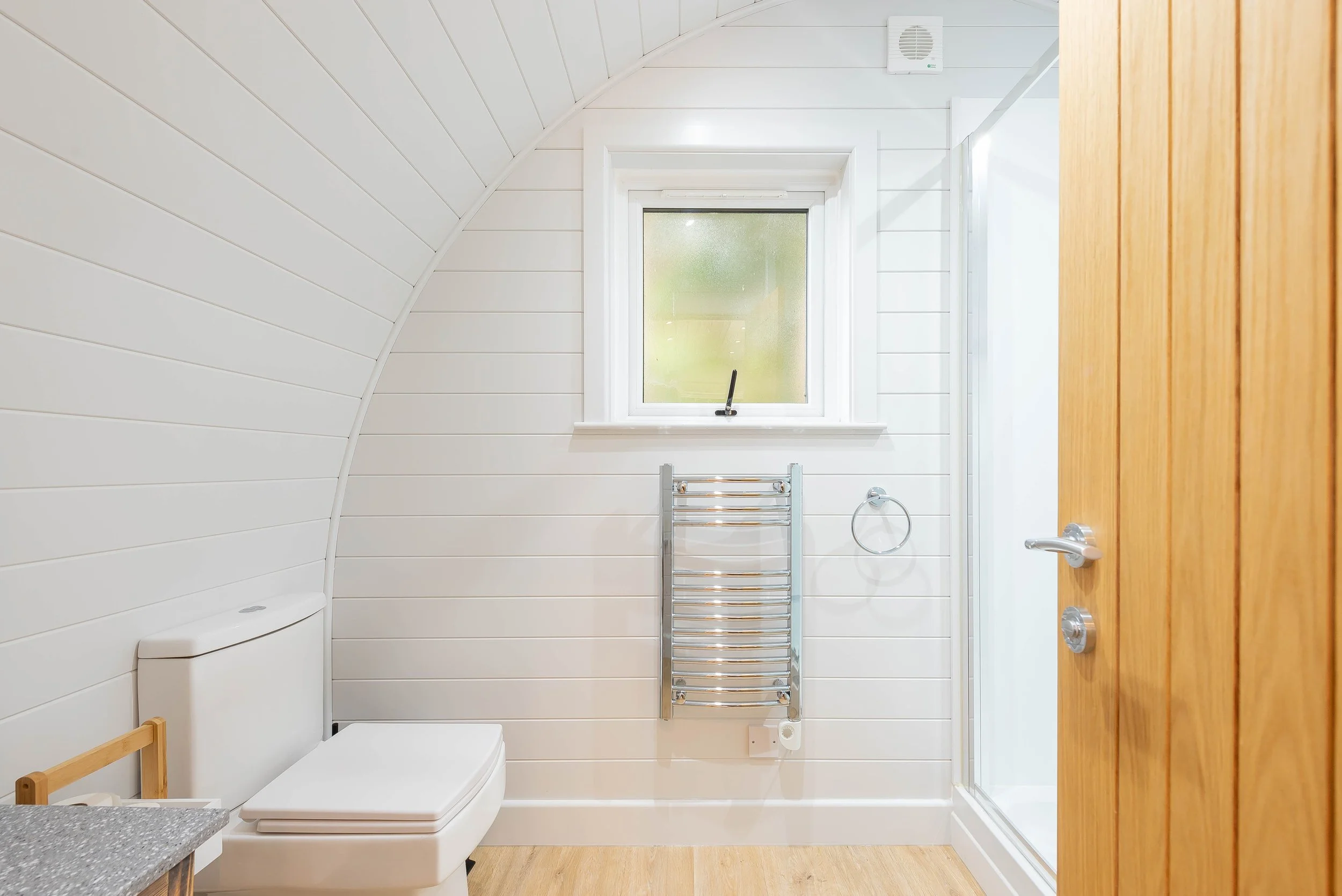A bright, modern bathroom with white wooden paneled walls and ceiling, a frosted window, a chrome towel warmer, a white toilet, and a glass-enclosed shower with a roll-up door. There is a wooden door on the right and a small wooden stool on the lower