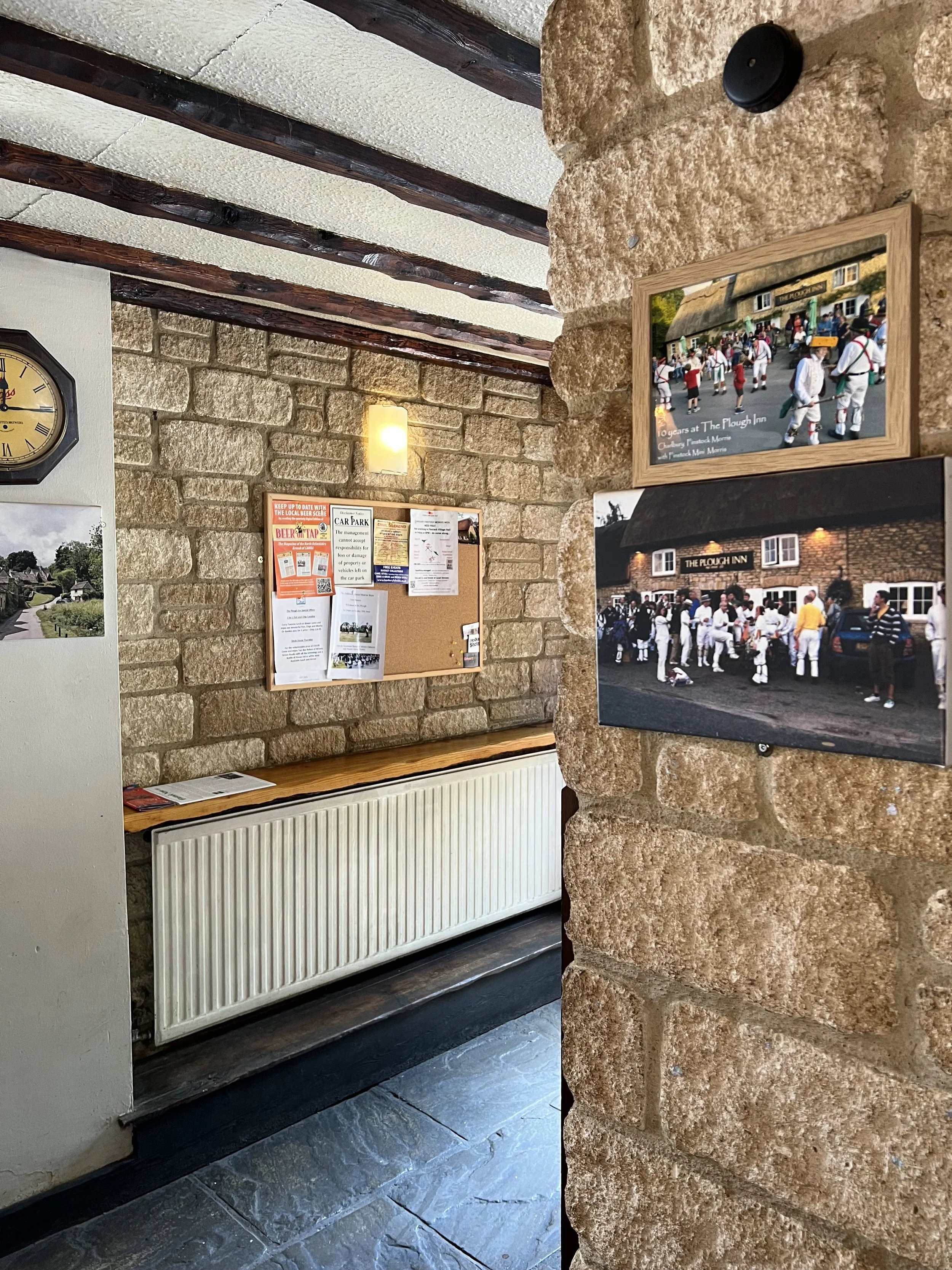 Interior of a rustic building with stone and brick walls, wooden ceiling beams, a wall clock, a radiator, and framed photographs of a historical reenactment outside The Plough Inn.