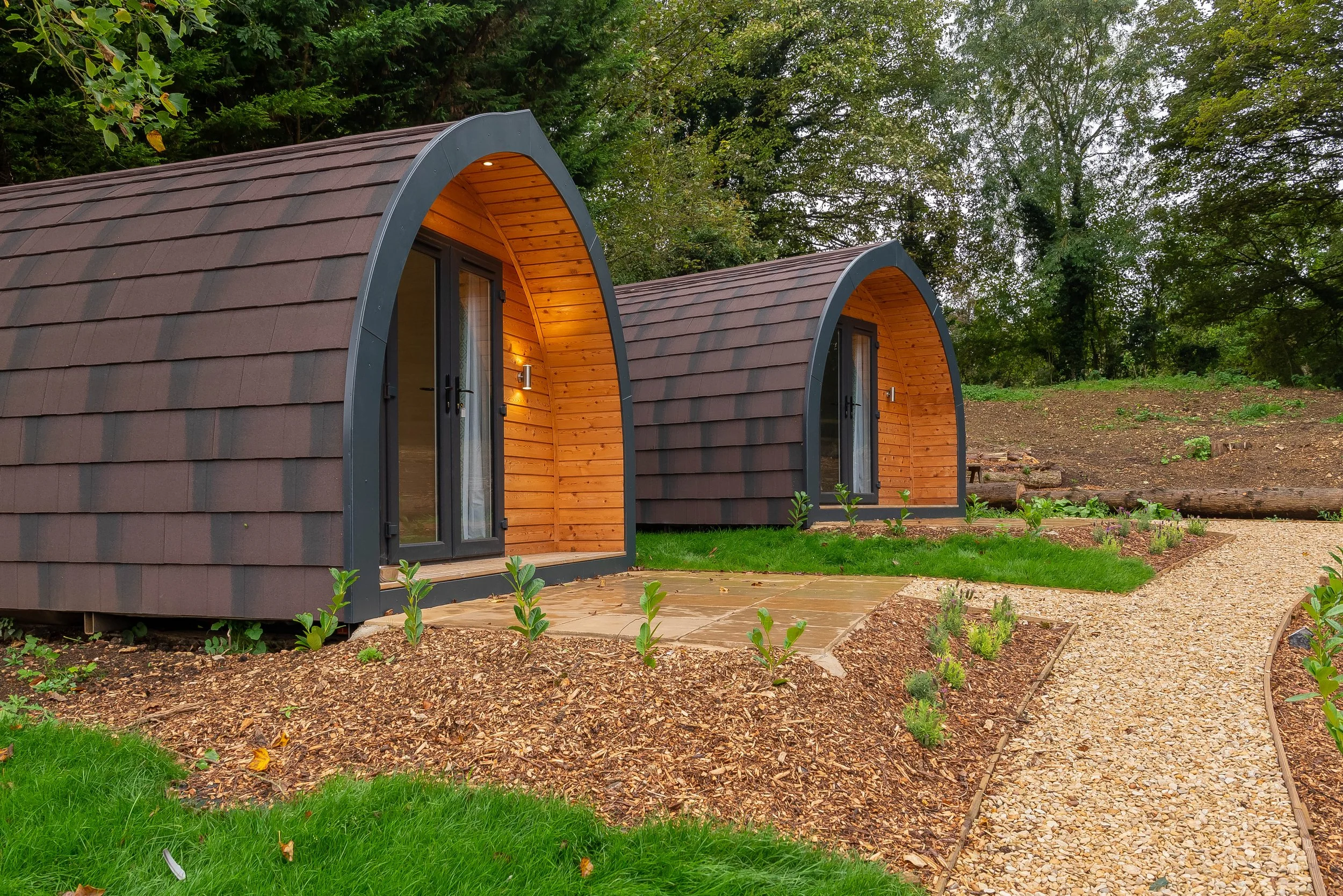 Two small, modern, egg-shaped cabins with shingled roofs and wooden interior entrances, situated in a lush green outdoor setting with trees and a gravel pathway.