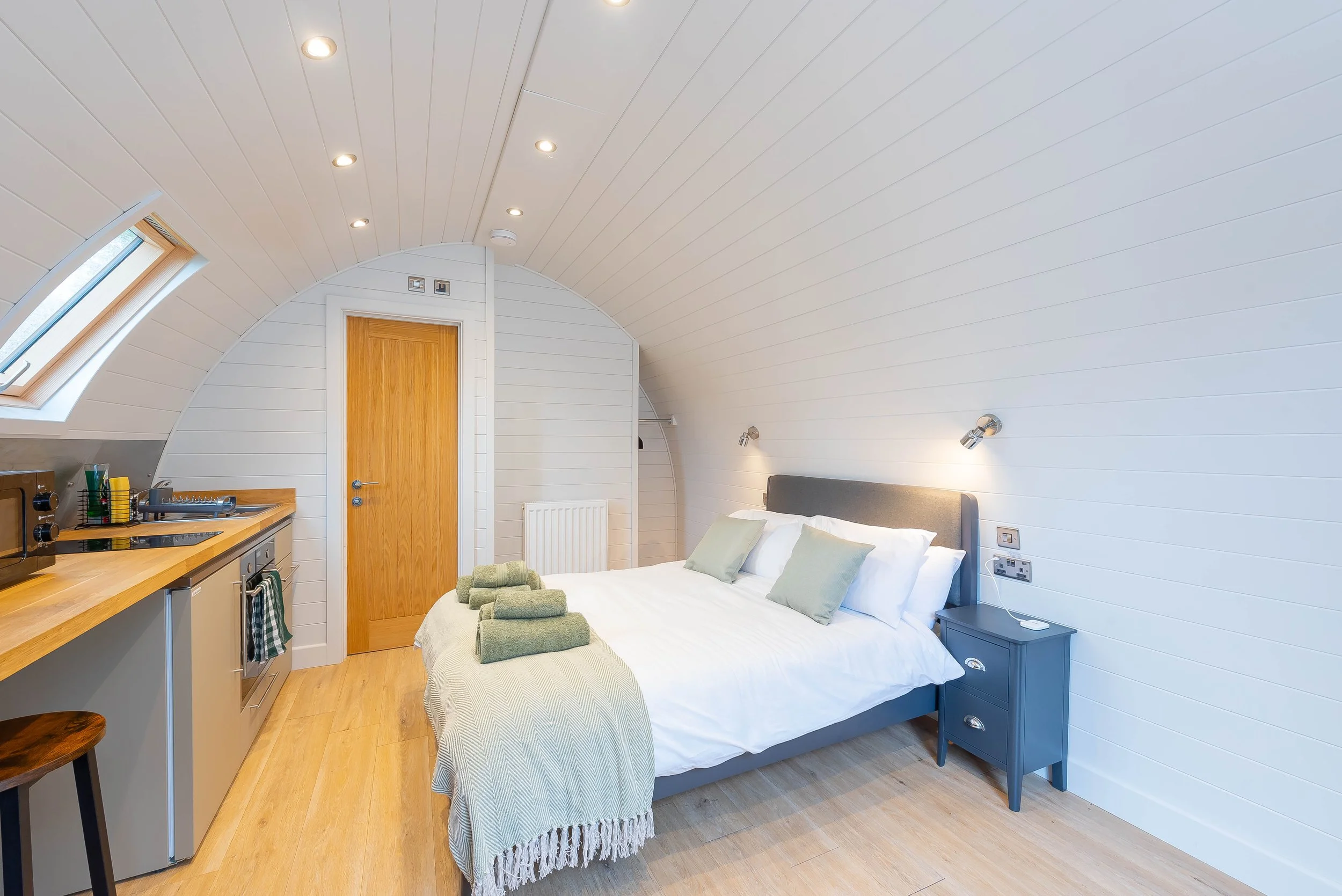 A cozy bedroom with white shiplap walls and ceiling, featuring recessed lighting and a skylight window. The bed has white bedding and light green cushions with neatly folded towels at the foot. There is a light blue bedside table and a small blue war