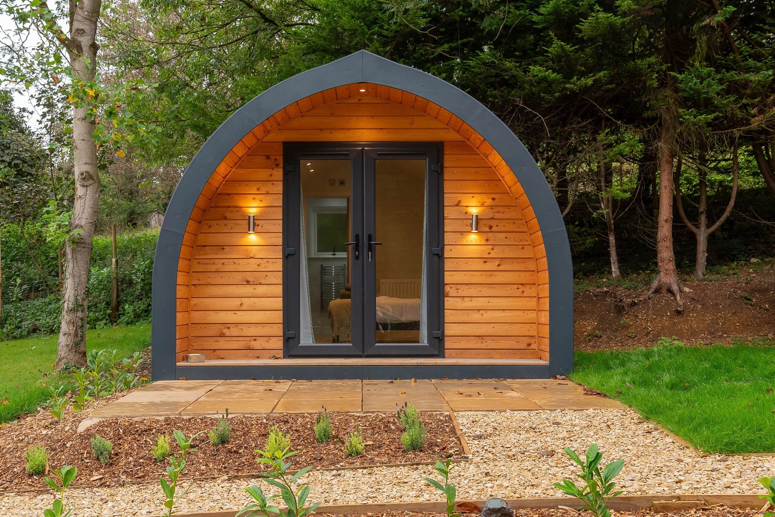 Small, modern wooden cabin with an arched roof, located in a wooded area, with glass double doors, outdoor lighting, a gravel and tile pathway, and garden plants in the foreground.