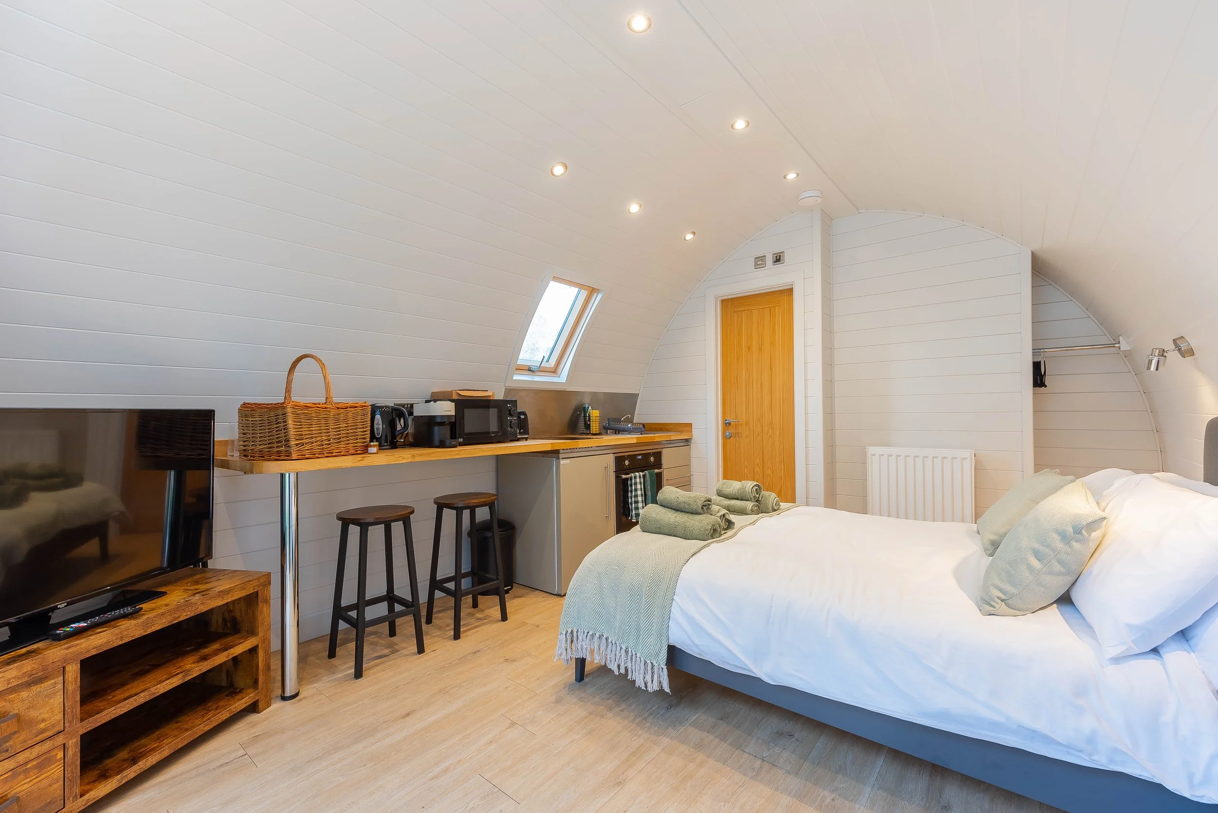 Interior of a bedroom with a curved white ceiling, illuminated by recessed lights, featuring a bed with white linens and a green throw blanket, a wooden door, a window, and a small kitchenette area with a microwave, coffee maker, and bar stools.