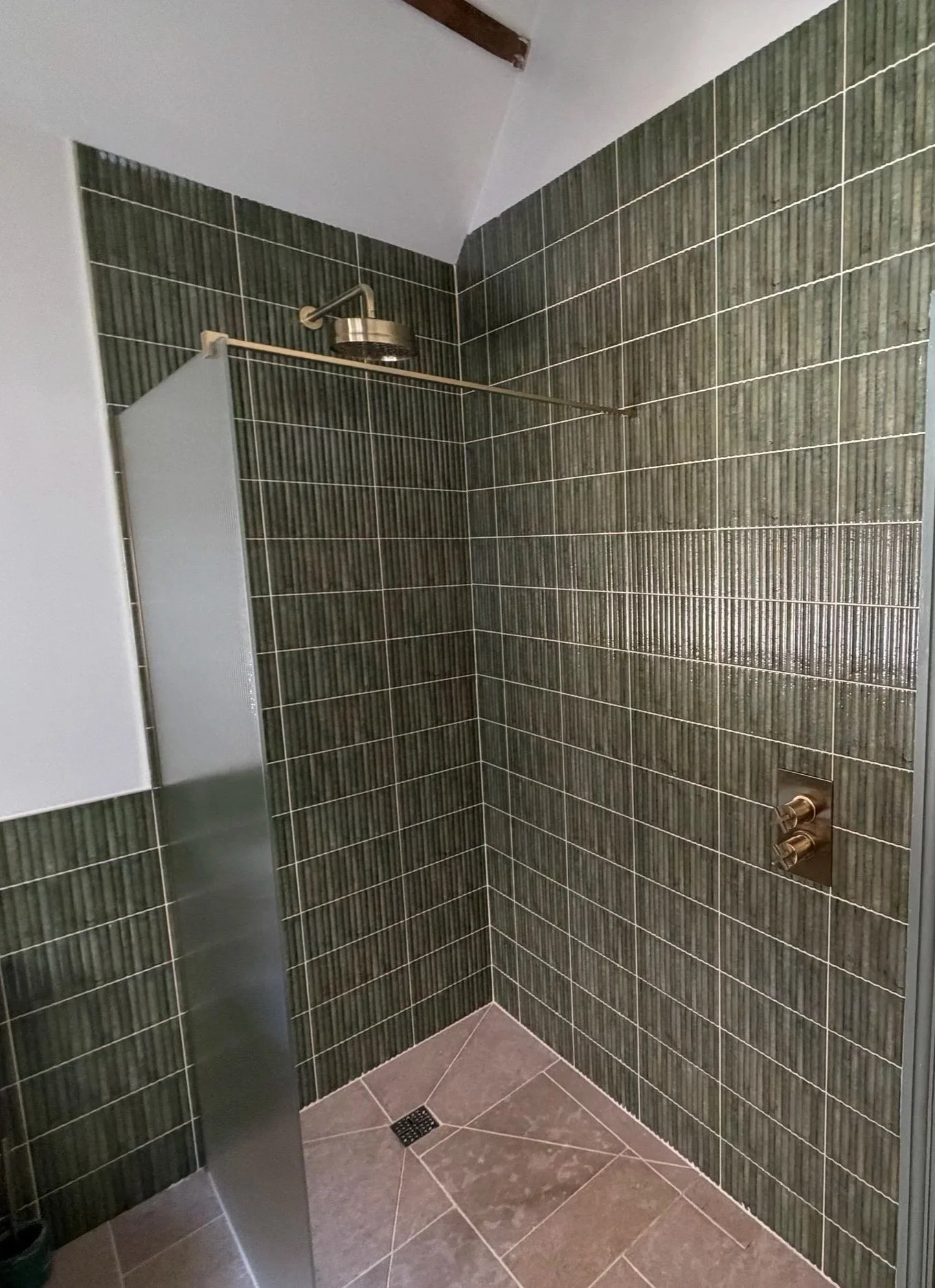A modern bathroom shower area with green vertically striped tiles. There is a handheld shower head mounted near the corner, and a small square drain on the gray tiled floor.