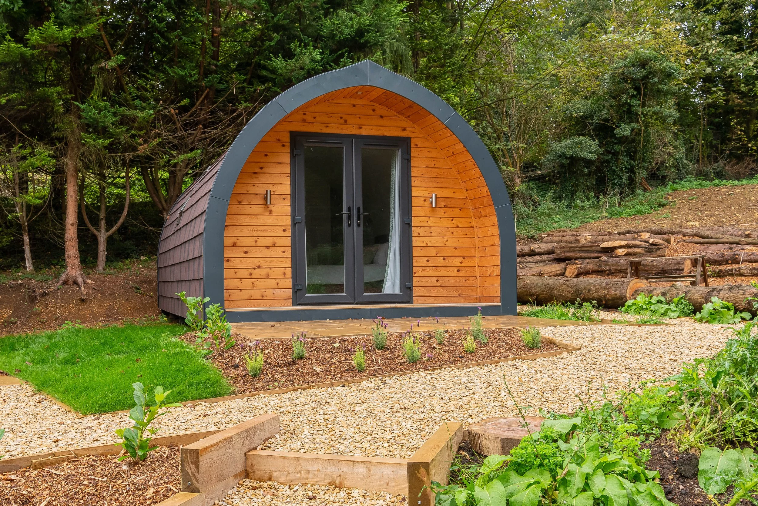 A small, modern, semi-circular wood cabin with a black door and windows, situated in a lush green forested area with a small flower garden and gravel pathways.