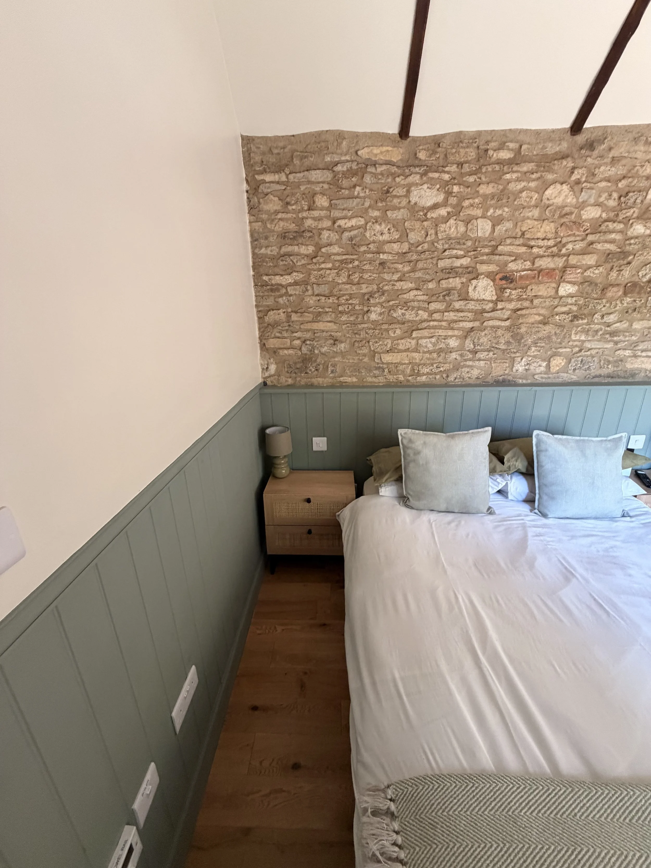 A bedroom corner with a bed, pillows, a wooden nightstand with a lamp, exposed brick wall, painted wood paneling in green, and hardwood flooring.