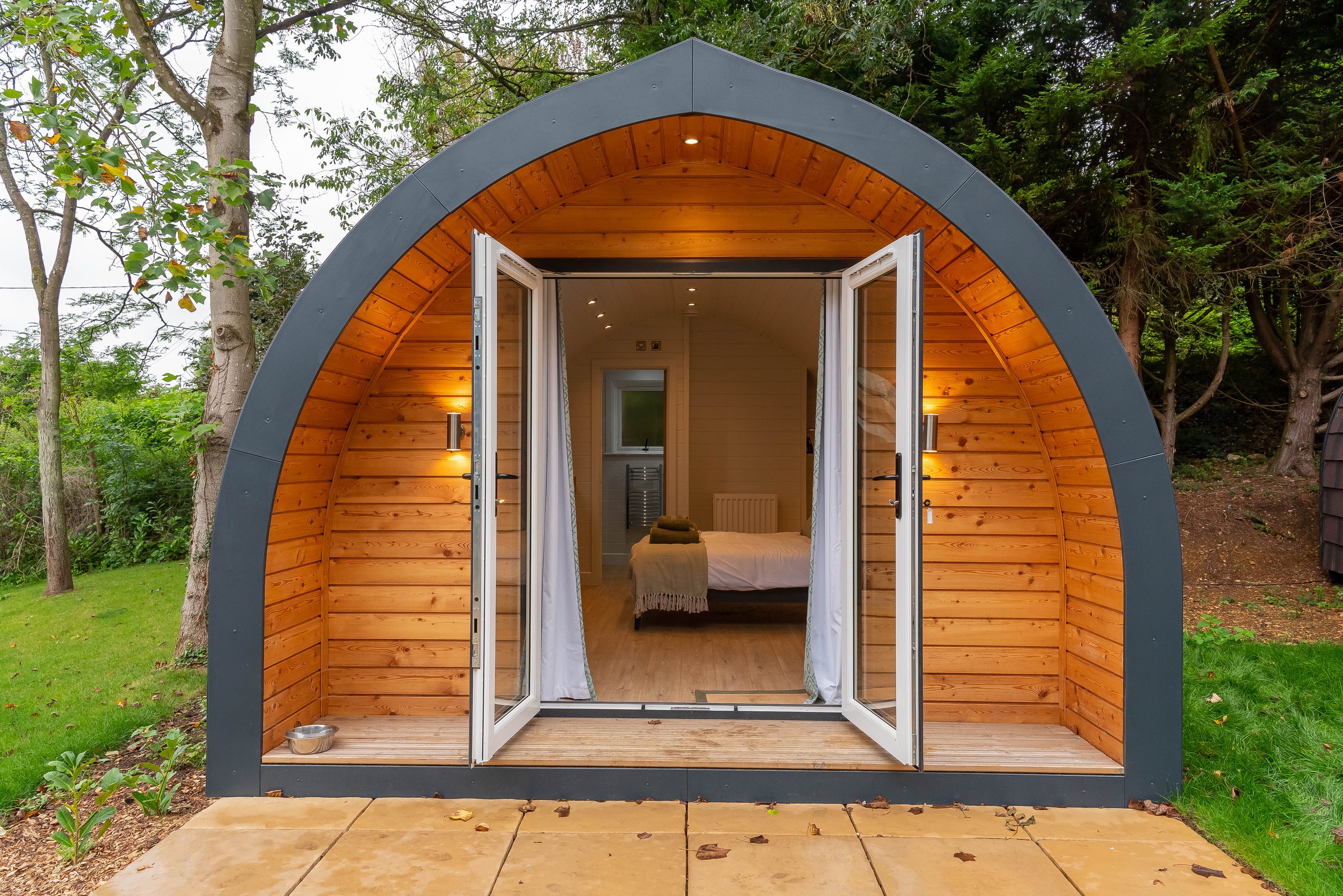 A small, modern, arched wooden cabin with glass doors open to reveal a cozy bedroom inside, surrounded by green trees and grass.