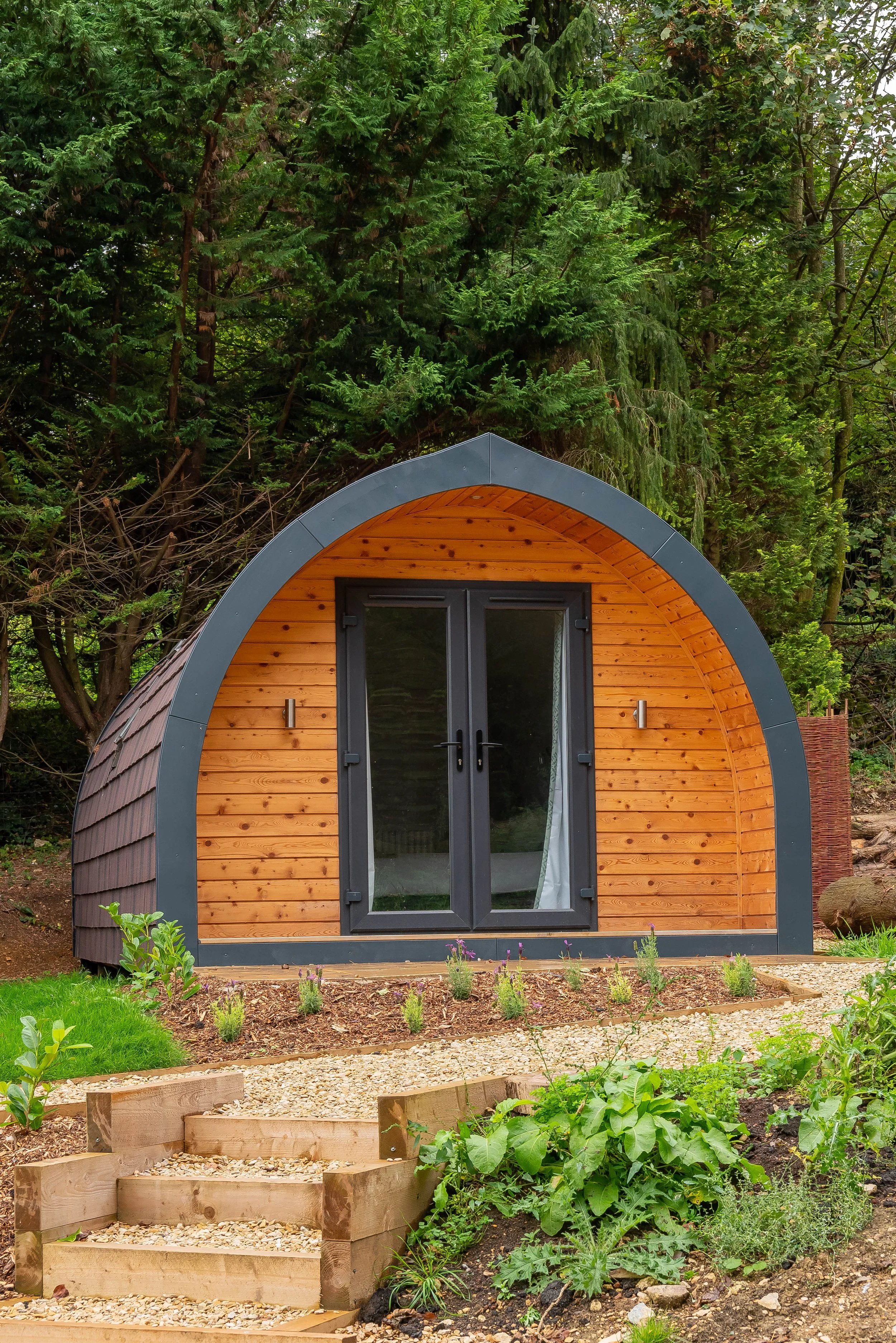 Small wooden and metal tiny house on wheels, set in a lush green garden with various plants and flowers, with a gravel pathway and steps leading to the door.