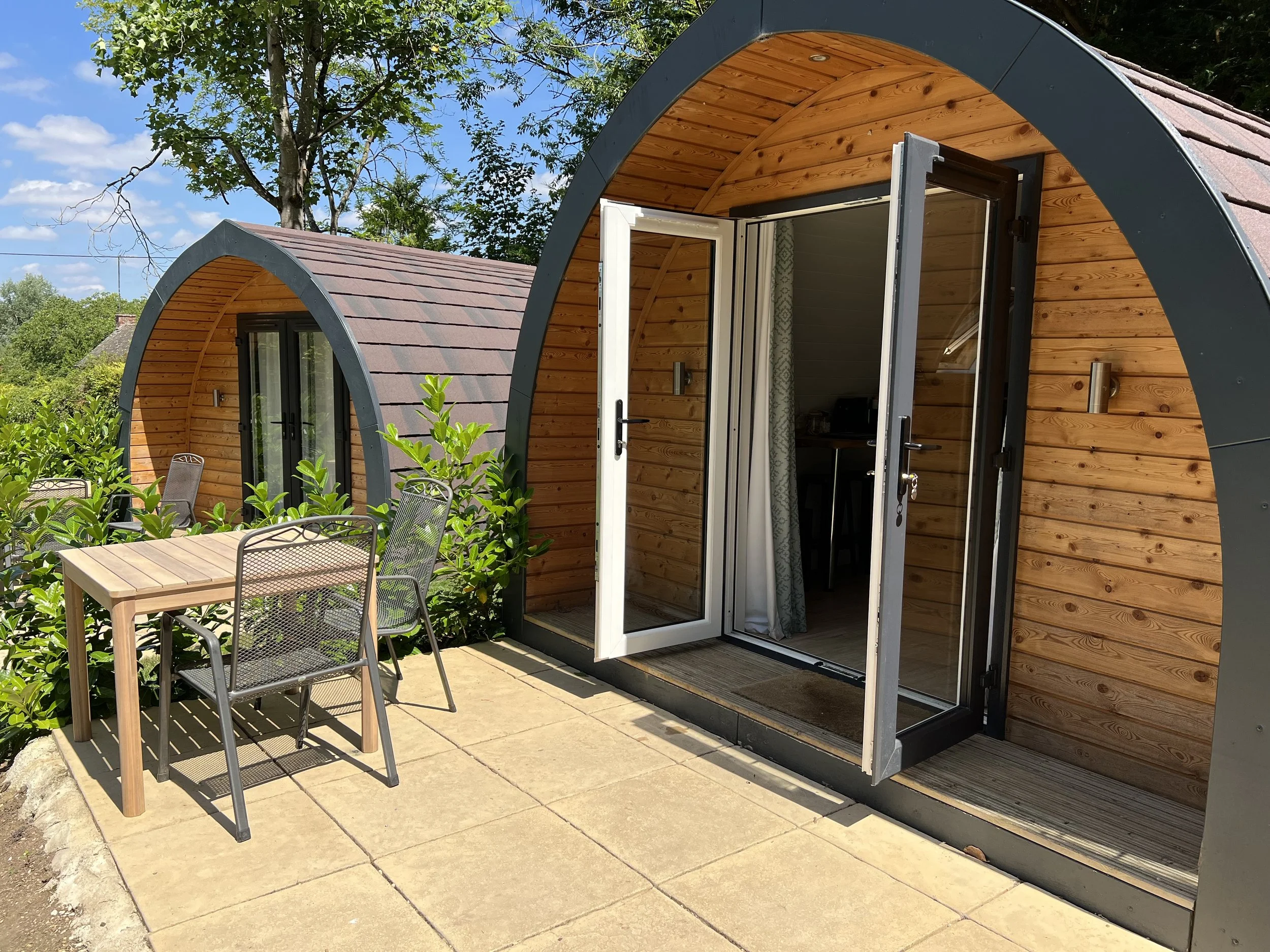 A small wooden cabin with an arched roof, located outdoors on a paved area. The cabin has an open door, letting a view of the interior. There is outdoor furniture, including a wooden table and metal chairs, nearby. Green bushes and trees surround the area, with a blue sky and some clouds overhead.