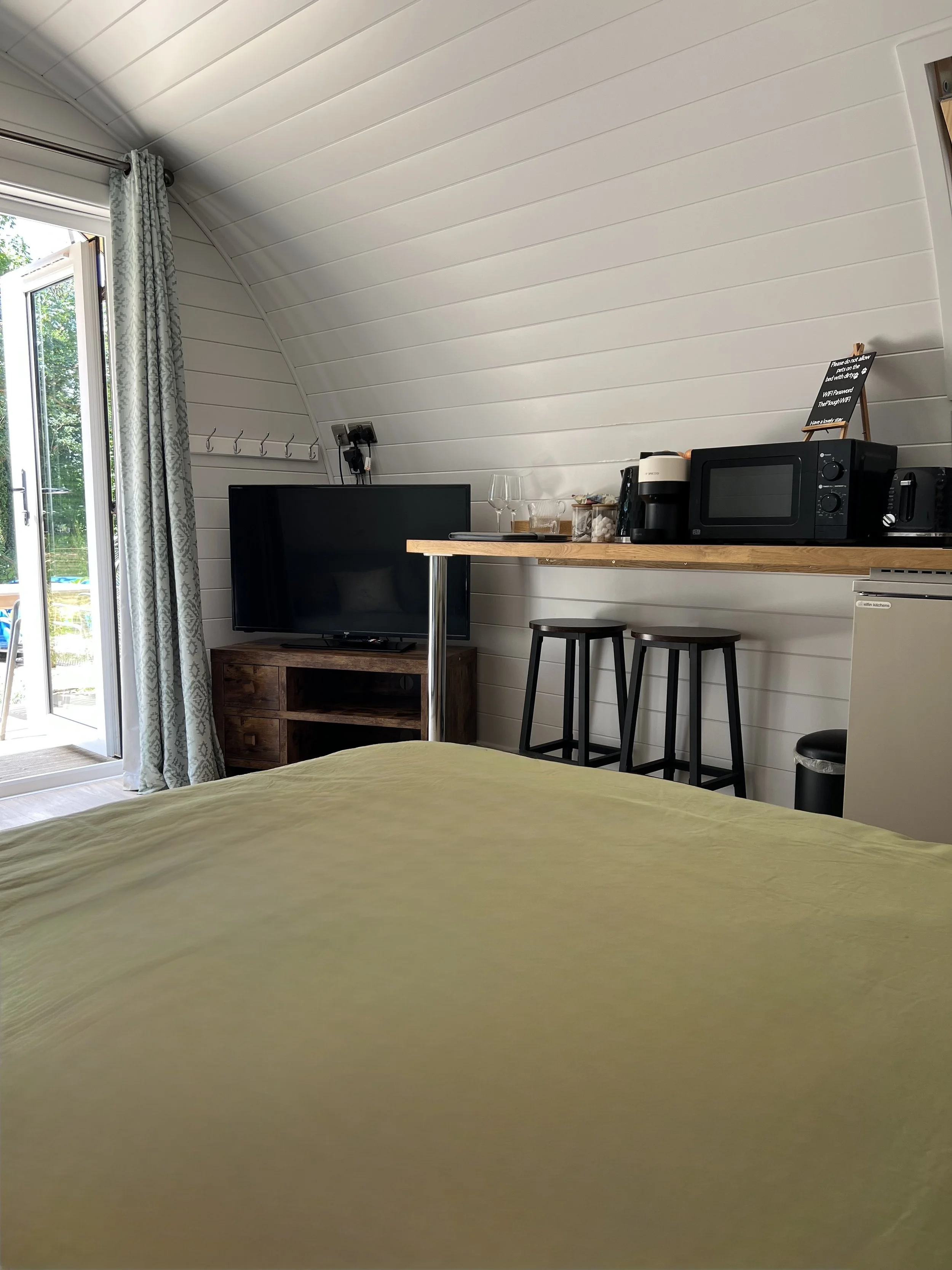 View of a cozy room with a yellow bedspread, a flat-screen TV on a wooden stand, a high wooden counter with bar stools, a microwave, coffee maker, wine glasses, and a small refrigerator, with a sliding glass door and curtains leading to a garden area.