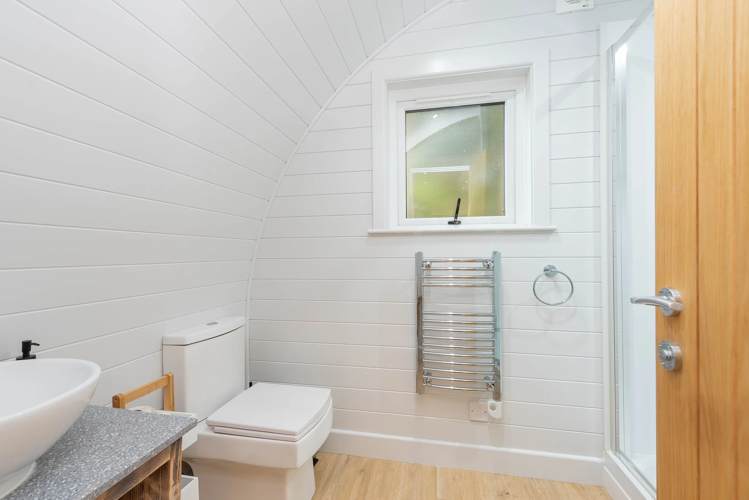 A modern bathroom with white shiplap walls, a small window, and a wooden door. It features a corner shower with a glass door, a wall-mounted towel heater, a toilet, and a countertop with a vessel sink.
