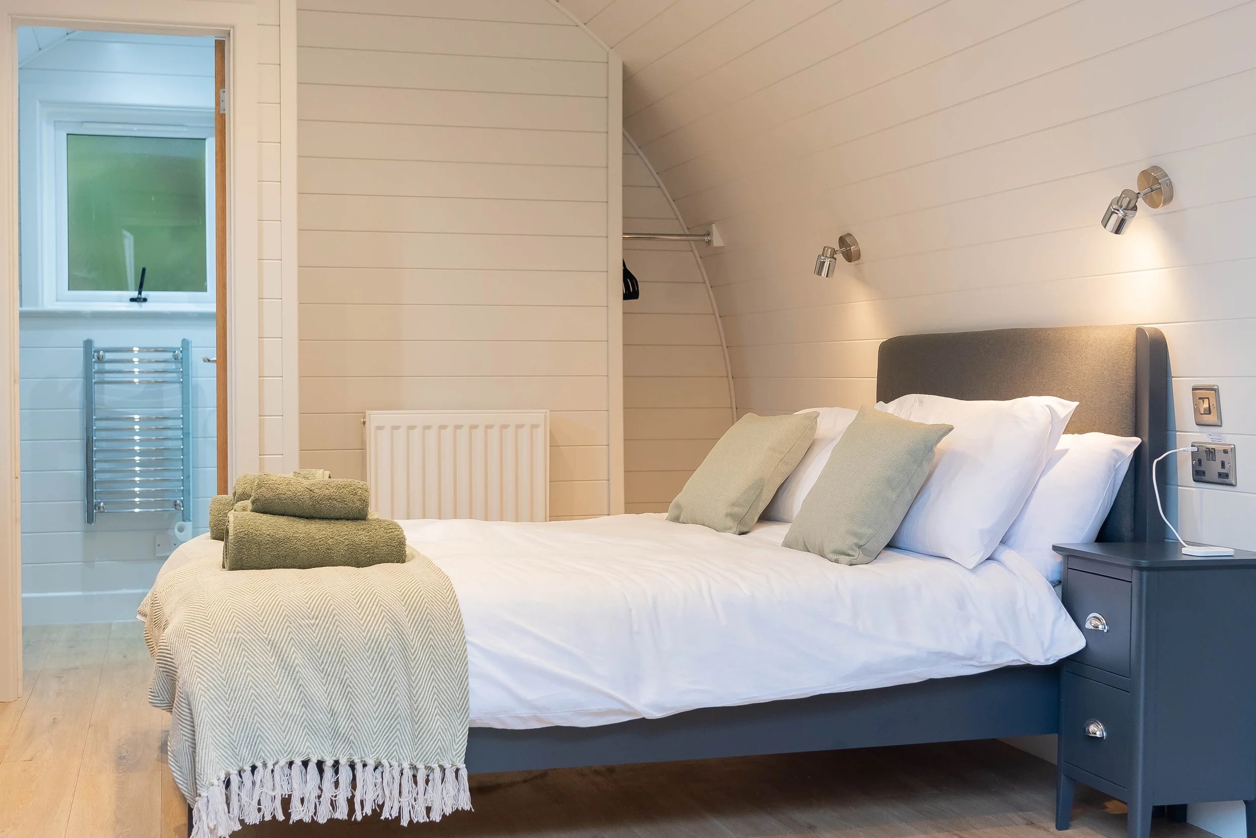 A bedroom with a double bed, gray headboard, white bedding, green pillows, and a beige throw blanket. There are rolled green towels on the bed. A nightstand with a lamp, electrical outlets, and a remote is beside the bed. The wall behind the bed is c