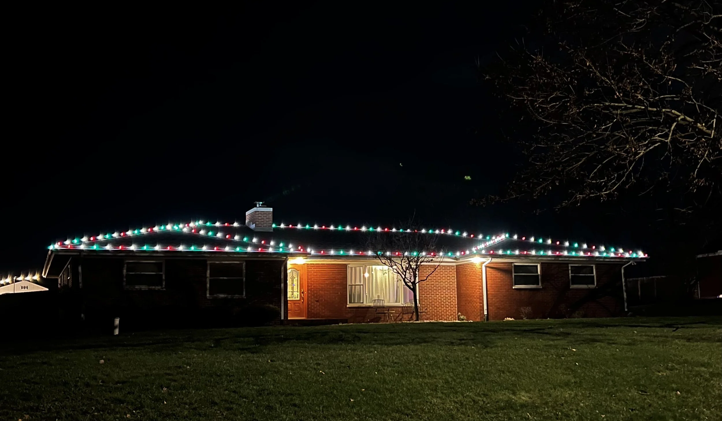 A brick house decorated with Christmas lights along the roofline at night. There is a tree in front and a window with curtains.