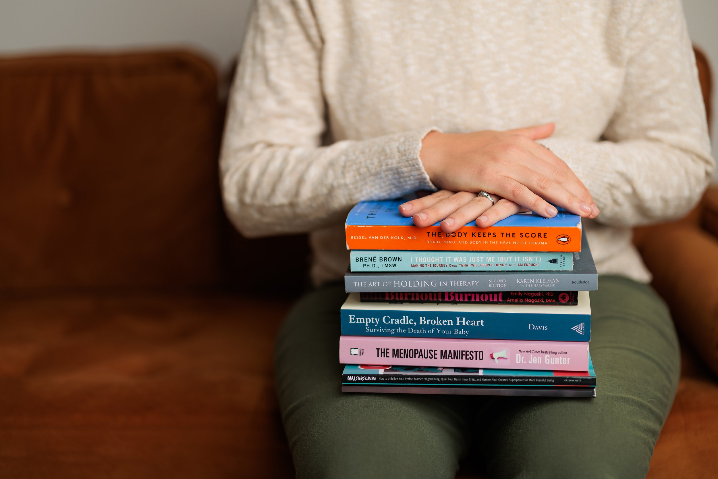 A person wearing a cream-colored sweater is sitting on a green surface with a stack of ten books on their lap. The books cover topics like trauma, therapy, menopause, and surviving loss.