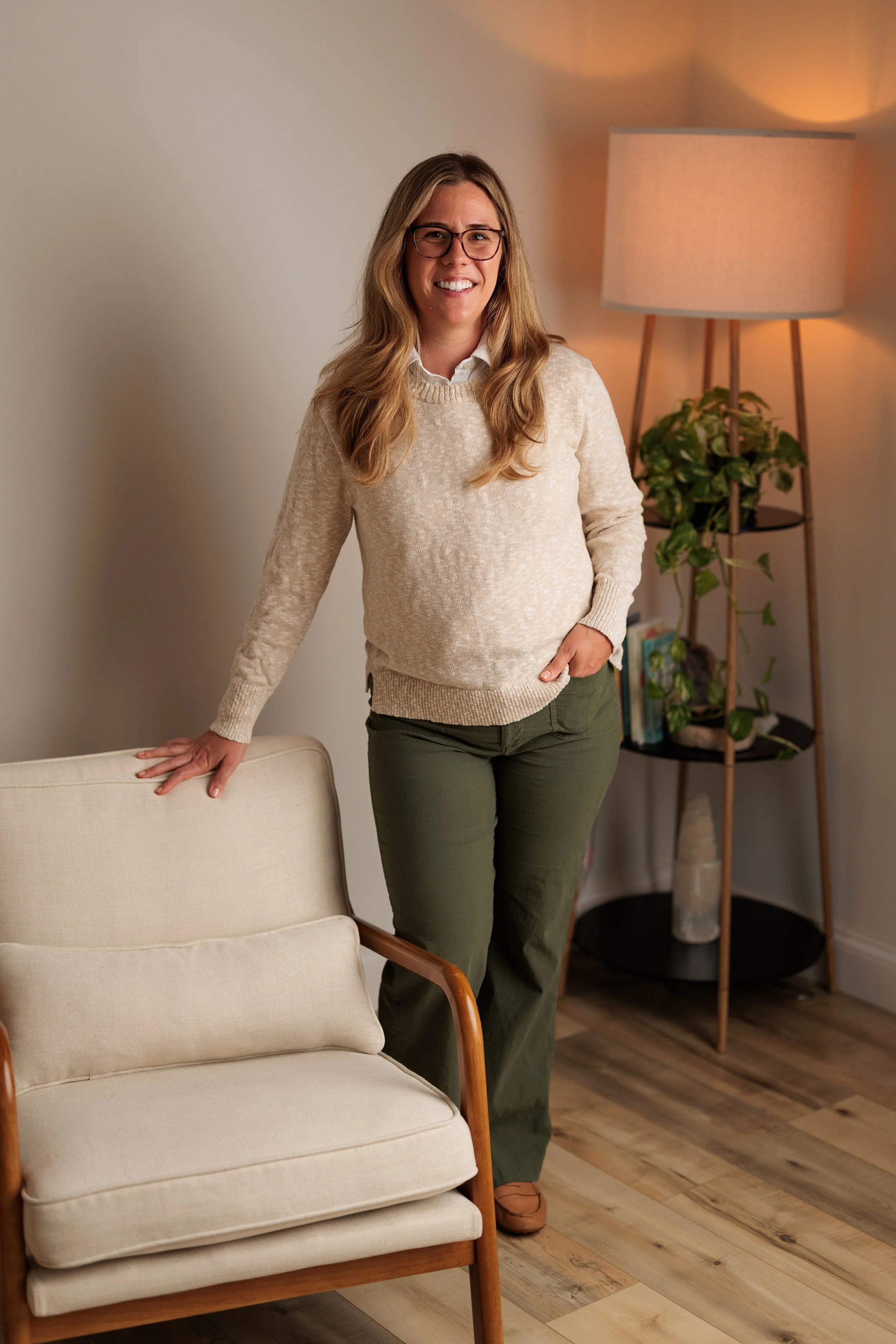 A woman with long blonde hair, glasses, wearing a beige sweater and green pants, leaning against a cream-colored couch in a living room with a wooden floor, a tall floor lamp, and a bookshelf with plants and books.