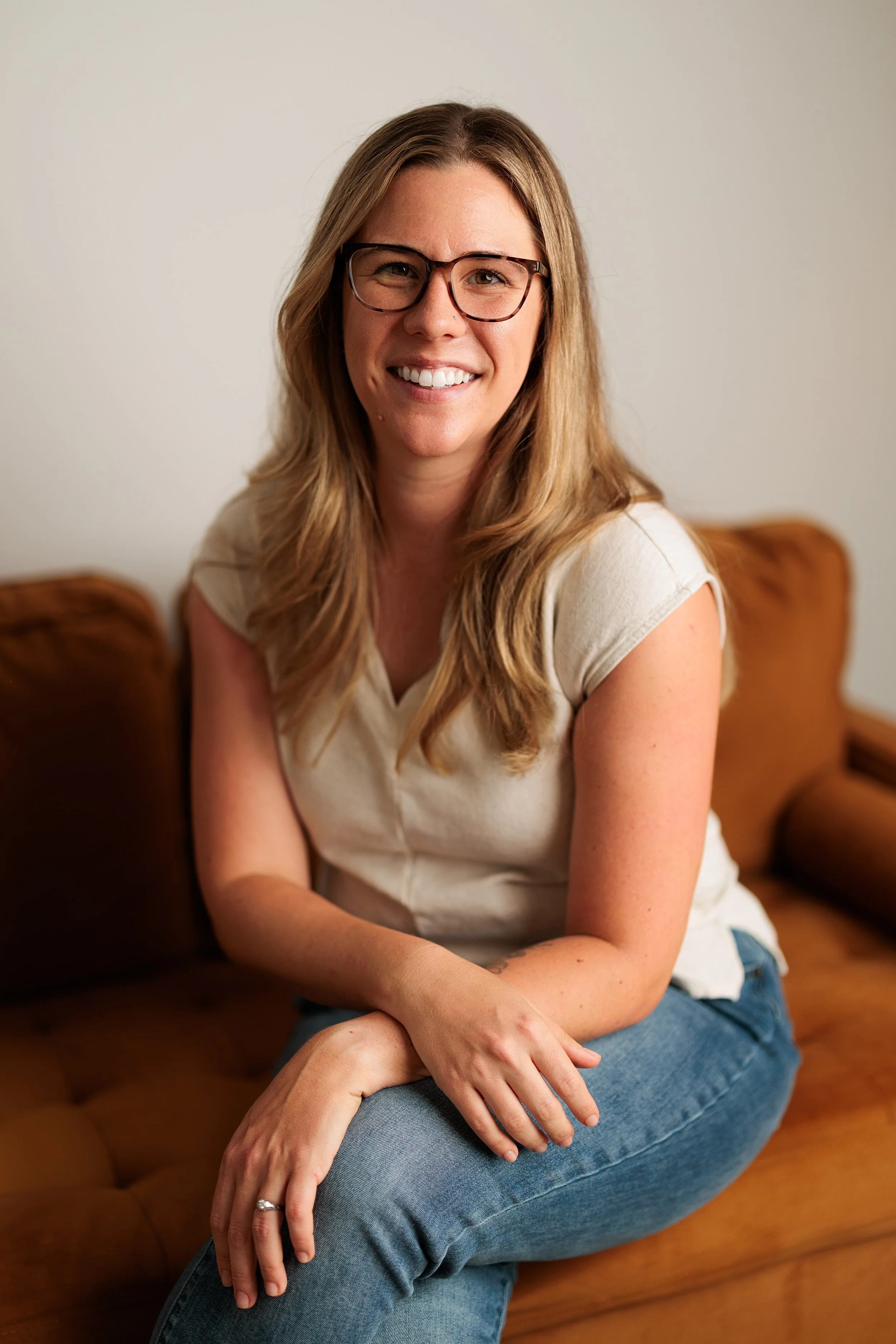 A woman with long blonde hair, glasses, and a white t-shirt sitting on a brown leather couch, smiling with her arms crossed, in a bright room with a neutral background.