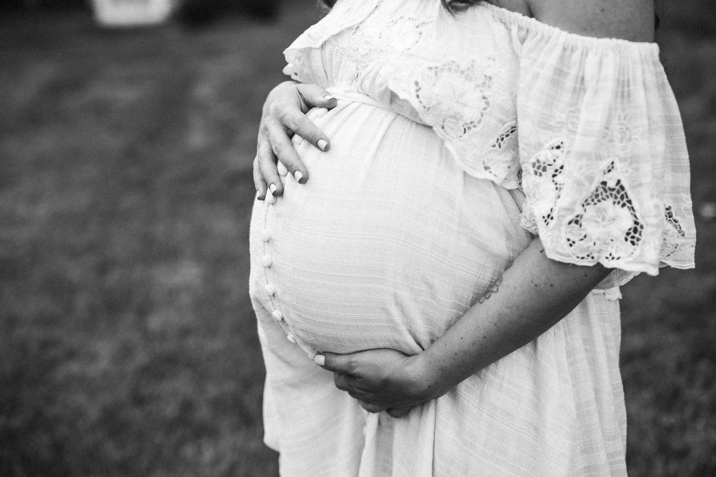 Close-up of pregnant woman wearing a white lace top and a flowing skirt, gently holding her belly with both hands, outdoors.
