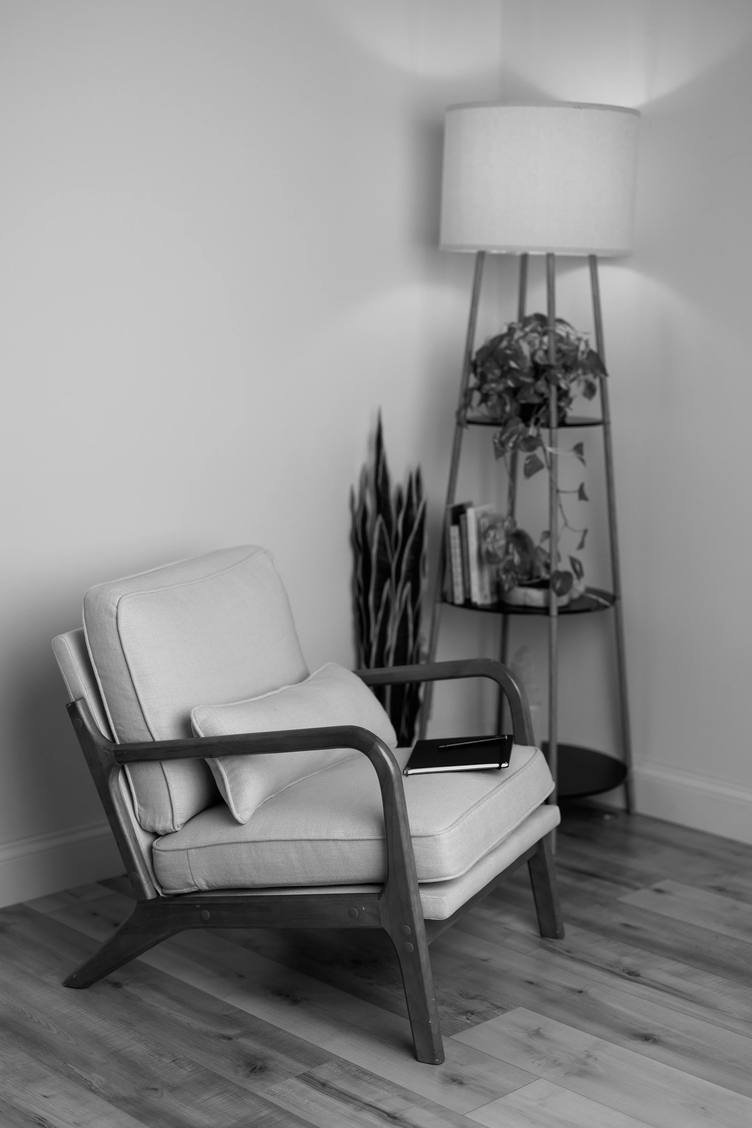 A mid-century modern armchair with a wooden frame and light-colored cushions, placed in a room with hardwood flooring. There is a tablet on the chair's seat, and behind it, a tall lamp with a fabric shade stands next to a bookshelf with books and a potted plant.