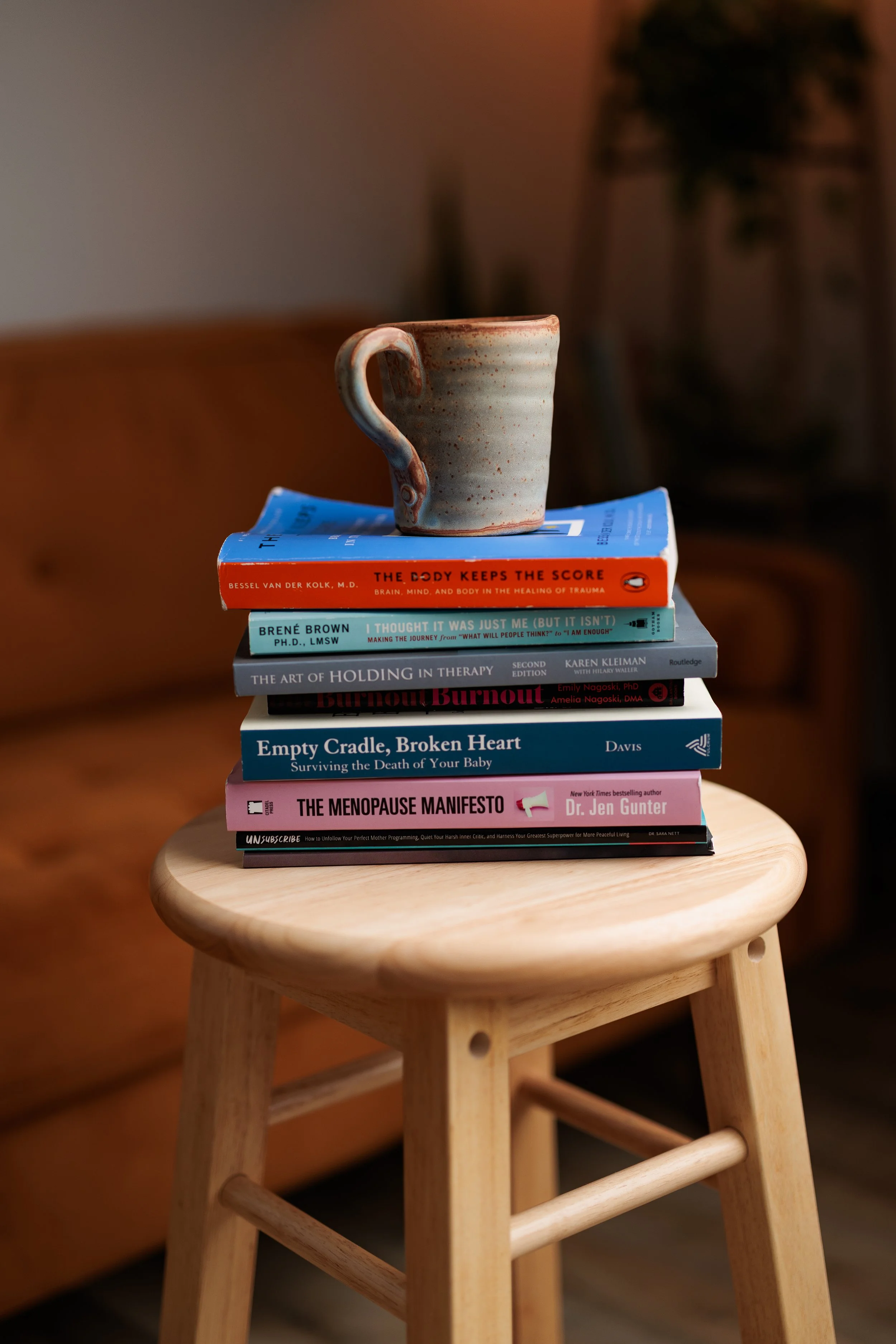 A small wooden stool with a stack of books on top, and a ceramic mug placed on the books. The background is blurred and dark, with a warm, cozy atmosphere.