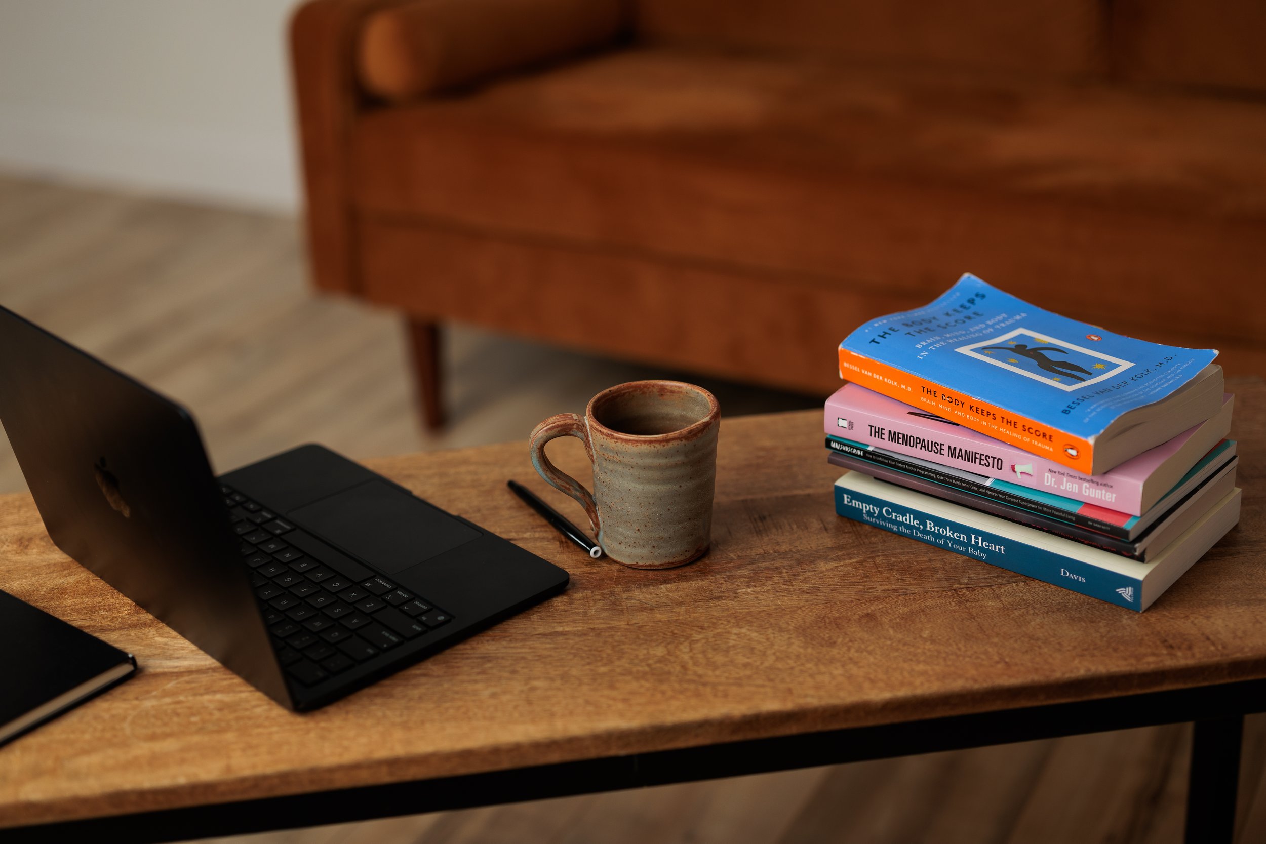 A wooden desk with a black laptop computer, a black notebook, a pen, a coffee mug, and a stack of seven books with colorful covers.