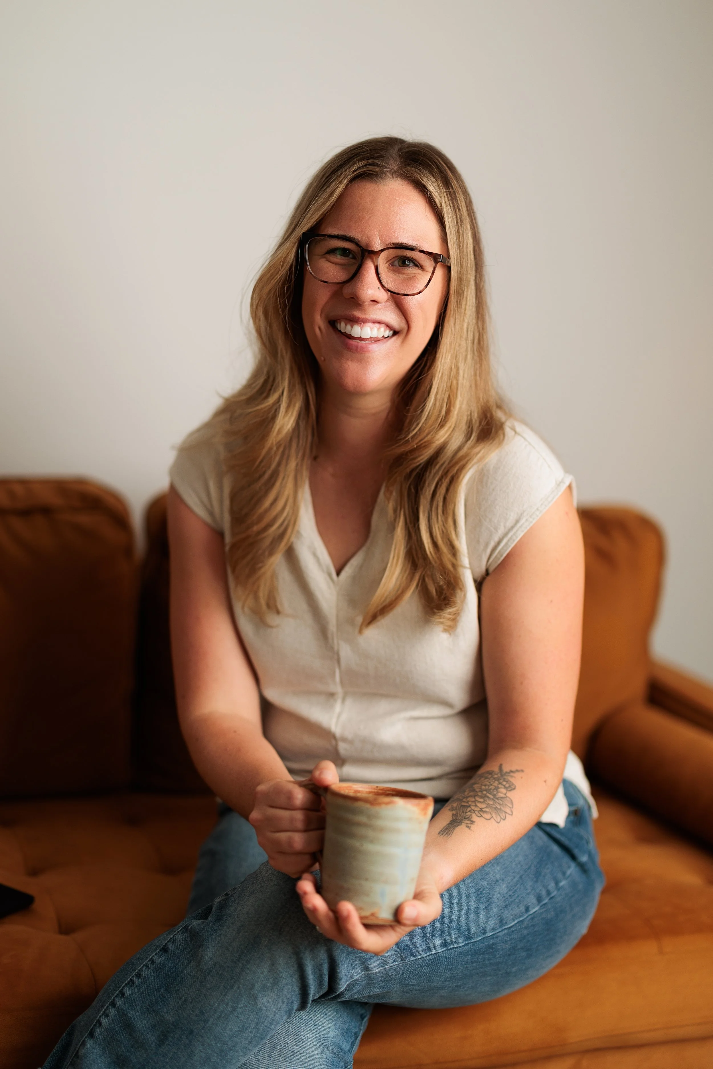 A woman with glasses and long wavy hair sitting on a brown leather couch, smiling and holding a ceramic mug, with a plain light-colored wall in the background.