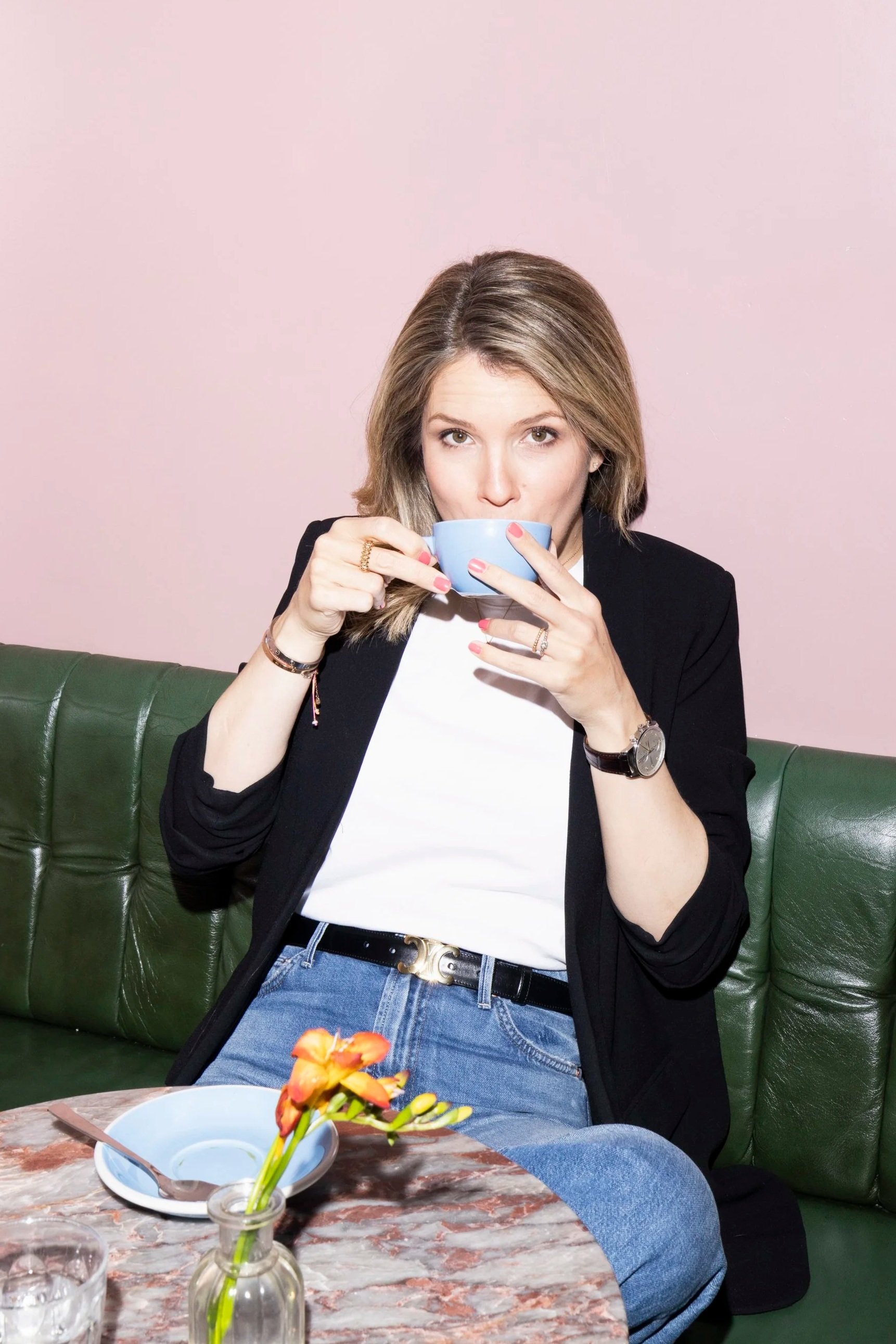 Eine junge Frau mit braunen, gestuften Haaren trinkt aus einer hellblauen Tasse, sitzt auf einer grünen Ledercouch vor einem rosa Hintergrund, trägt eine schwarze Jacke, weißen T-Shirt und jeans, trägt mehrere Ringe und eine Armbanduhr.