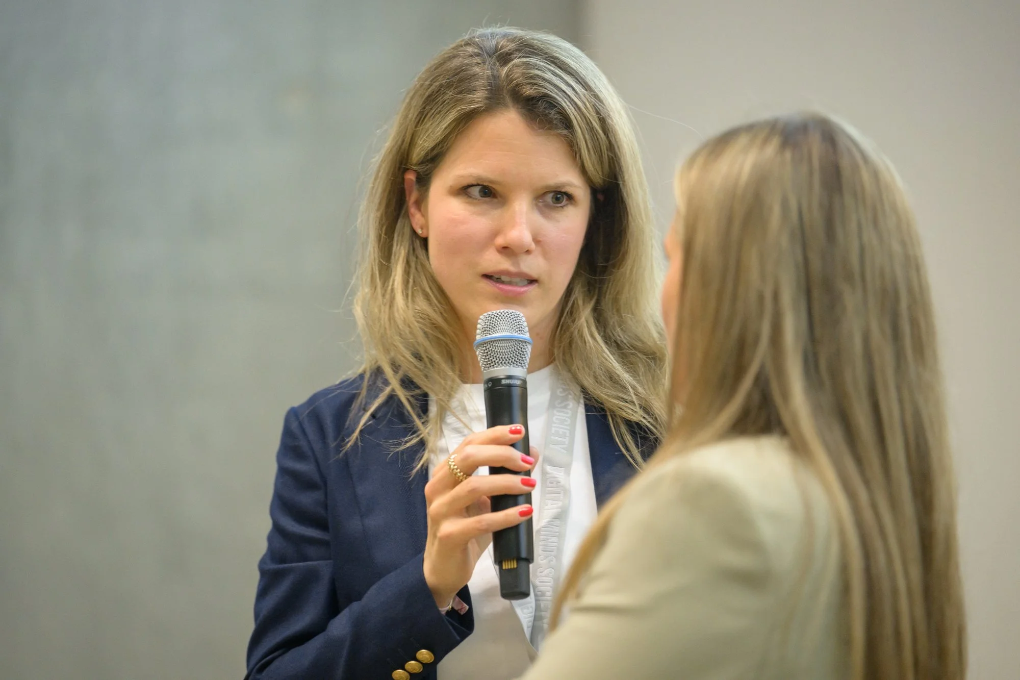 Eine Frau spricht mit einem Mikrofon auf einer Konferenz. Sie trägt einen dunklen Blazer und hat lange, blonde Haare. Im Vordergrund ist eine andere Person zu sehen, deren Rücken und blonde Haare sichtbar sind.