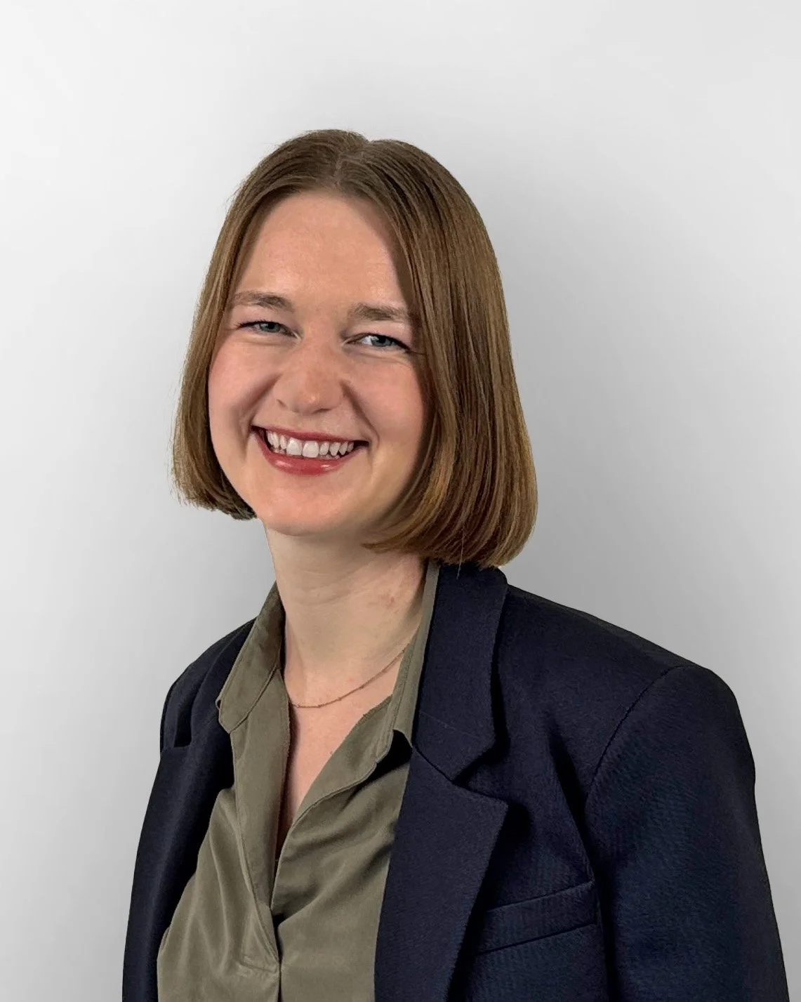 Porträt einer lächelnden jungen Frau mit mittellangen braunen Haaren, dunkler Blazer und beigefarbener Bluse vor einem weißen Hintergrund.