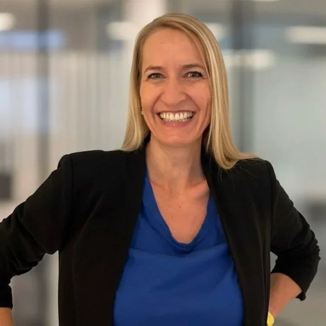 Lächelnde Frau mit blonden Haaren in einem schwarzen Blazer und blauer Bluse, stehend in einem modernen Büro.