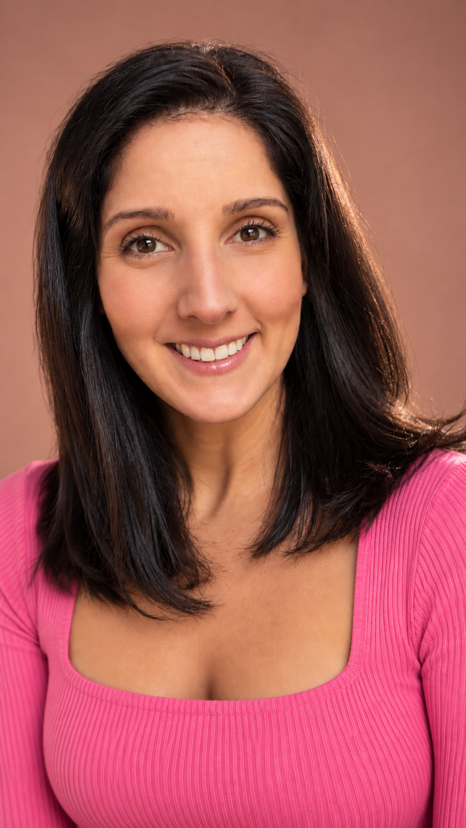 A woman with dark hair smiling, wearing a pink ribbed top, against a brown background.