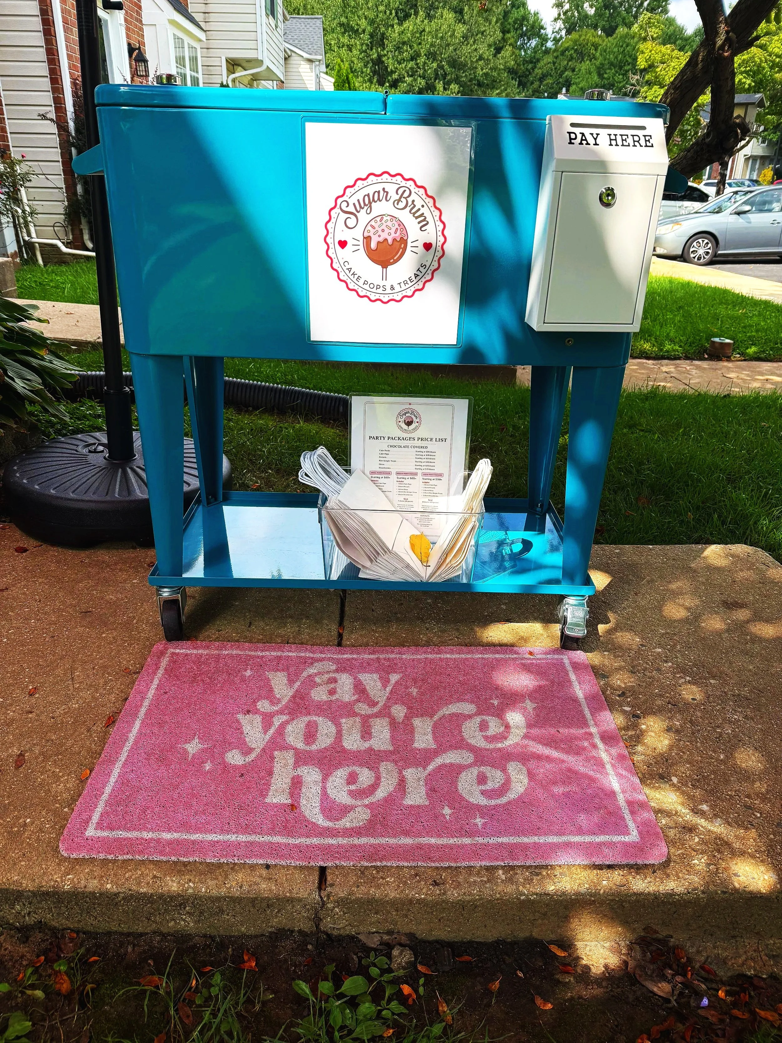 A blue outdoor cart with a pink mat in front reading 'yay you're here.' The cart displays a sign for 'Sugar Brim Cake Pops & Treats' and a small paper menu with napkins underneath.