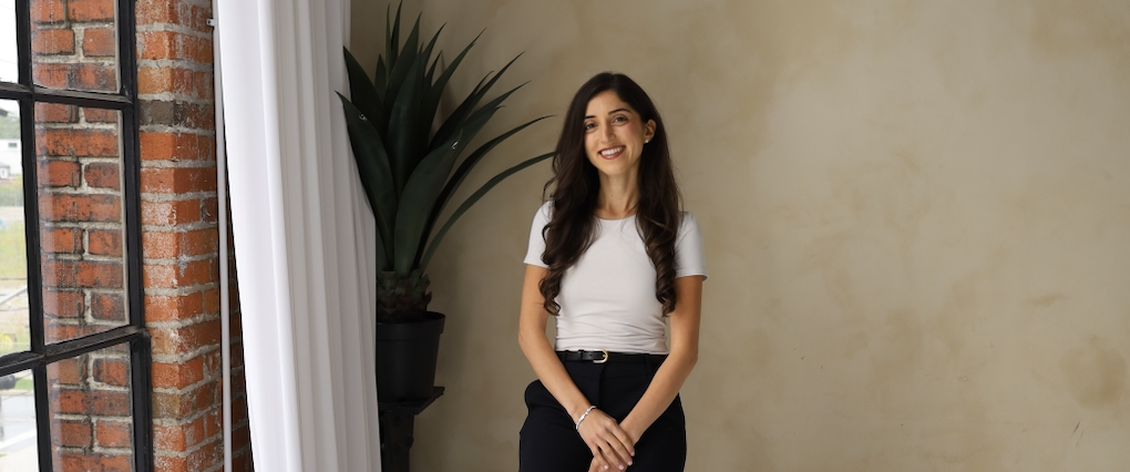 A woman with long dark hair, wearing a white t-shirt and black pants, is smiling and standing indoors next to a large green plant with a brick wall and window in the background.