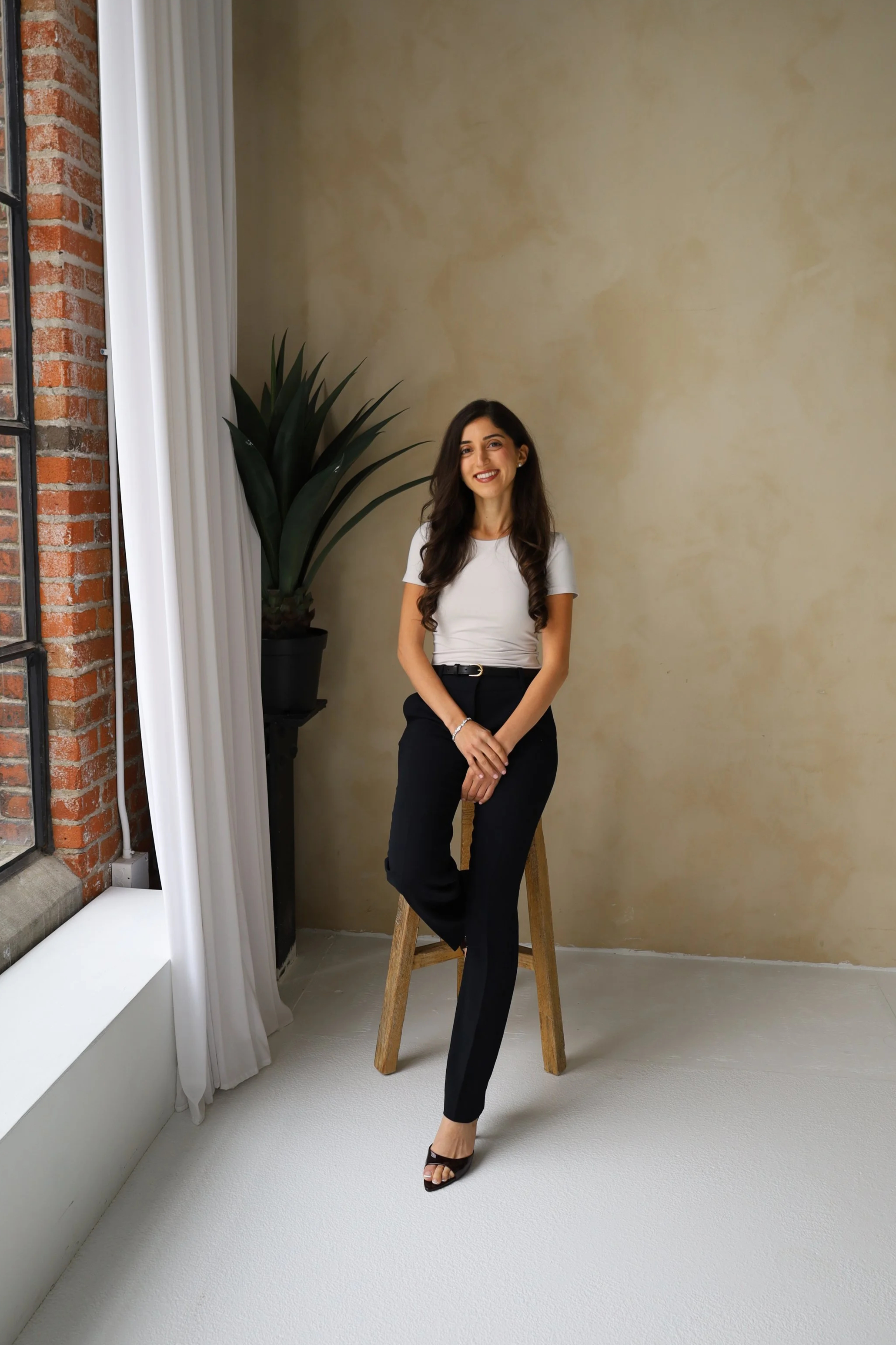 A woman with long dark hair, dressed in a white top and black pants, smiling and posing while standing on a wooden stool in front of a beige wall inside a room with a large window and white curtains.