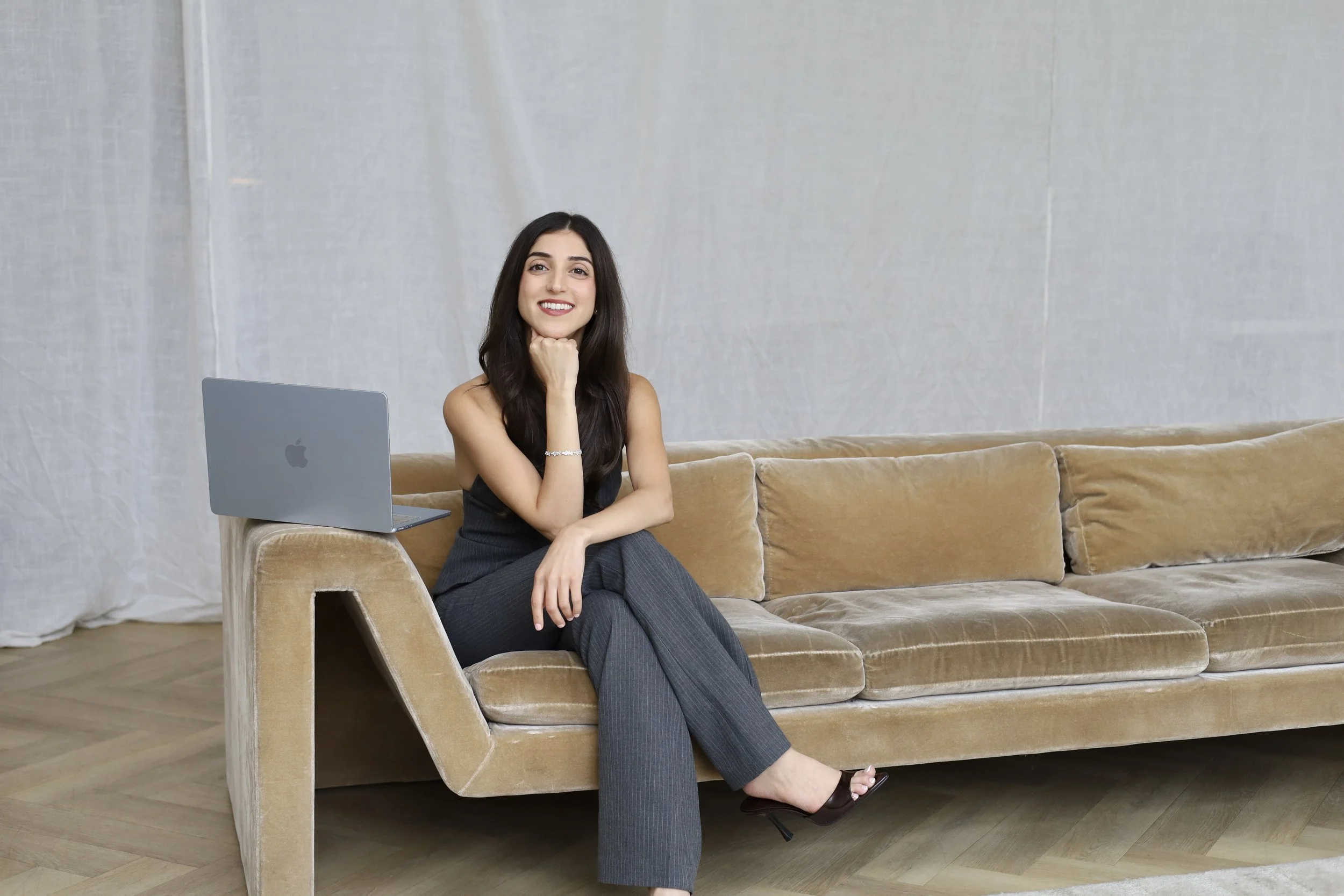 A woman sitting on a tan velvet couch with an open MacBook beside her, smiling, with her chin resting on her hand, in a minimalist room with a gray wall background.