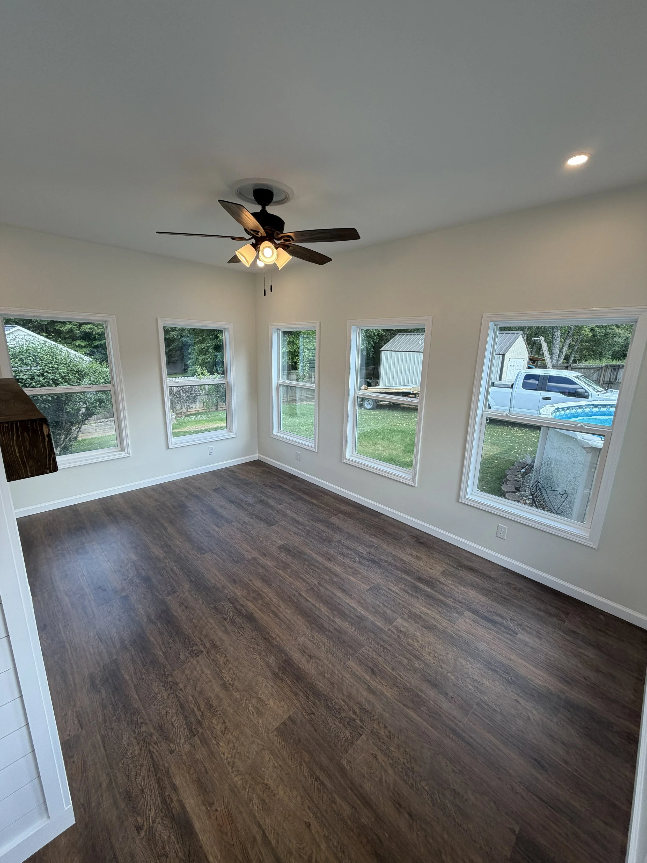 Living room with four large windows, a ceiling fan with light fixtures, and dark wood flooring. Outside, a white truck, a shed, and trees are visible.