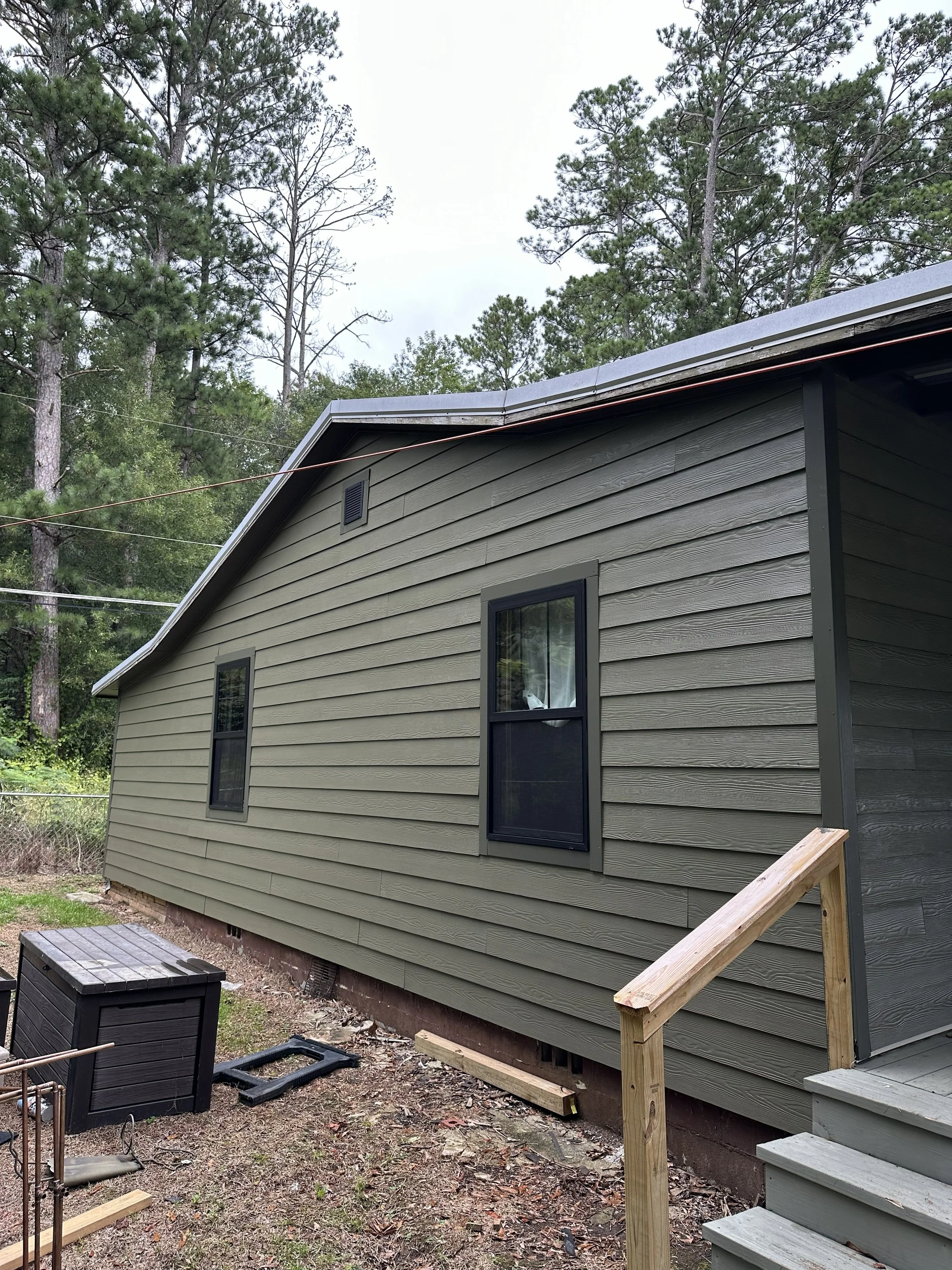 A house with green siding, black shutters, and a metal roof, with a wooden staircase and steps leading to a door. There are trees in the background and some construction materials on the ground.