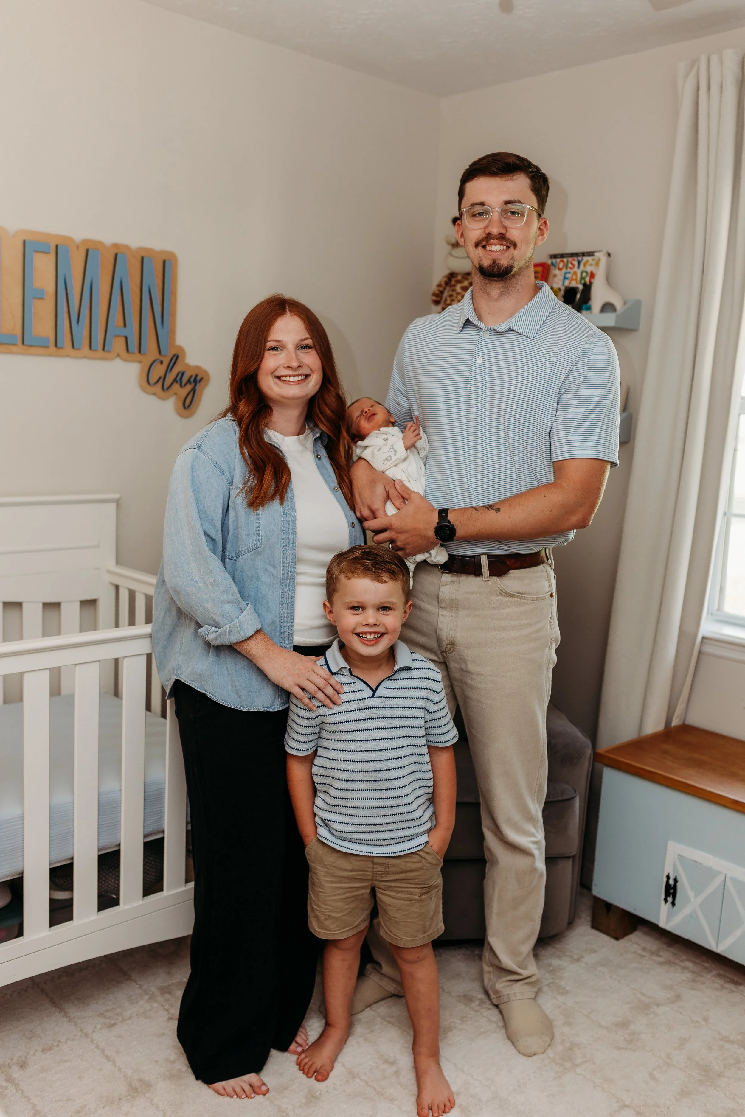 A happy family of five posing in a nursery. The family includes a woman with long red hair, a man with glasses, a toddler boy, an older boy, and a newborn baby. The woman is wearing a light denim shirt and black pants, the man is wearing a striped shirt and light-colored pants. The toddler is wearing a striped shirt and khaki shorts. Behind them, there is a sign that reads 'Gentleman Clay' and a framed picture with stuffed animals on a shelf. The room has a beige carpet, white crib, and curtains.