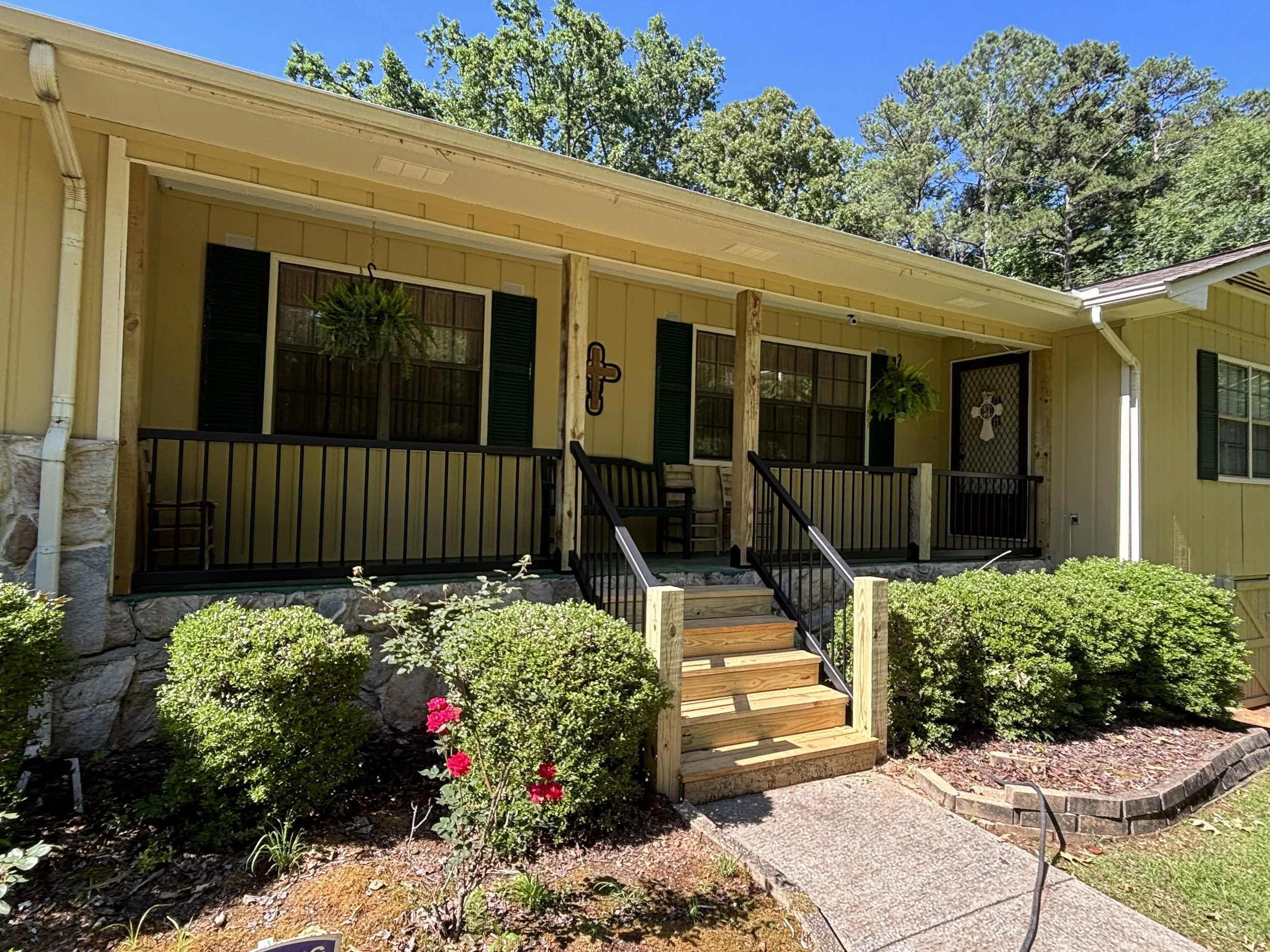 Yellow house with green shutters and a small front porch, surrounded by garden plants and flowers, with a blue sky and tall trees in the background.
