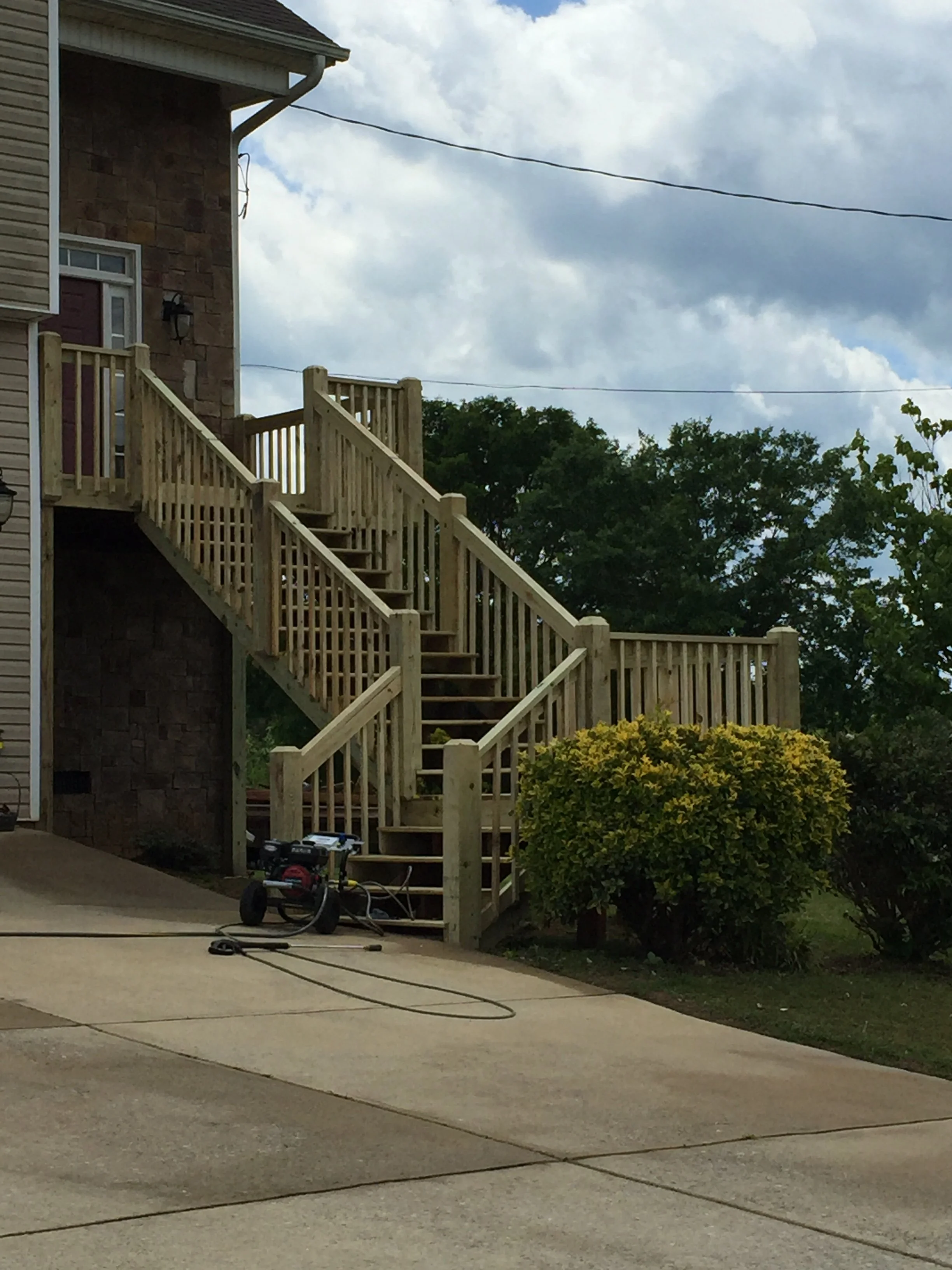 A wooden outdoor staircase attached to the side of a house, with construction tools and equipment on the concrete landing below, a bush with yellow-green leaves, green trees, cloudy sky, and overhead power lines.